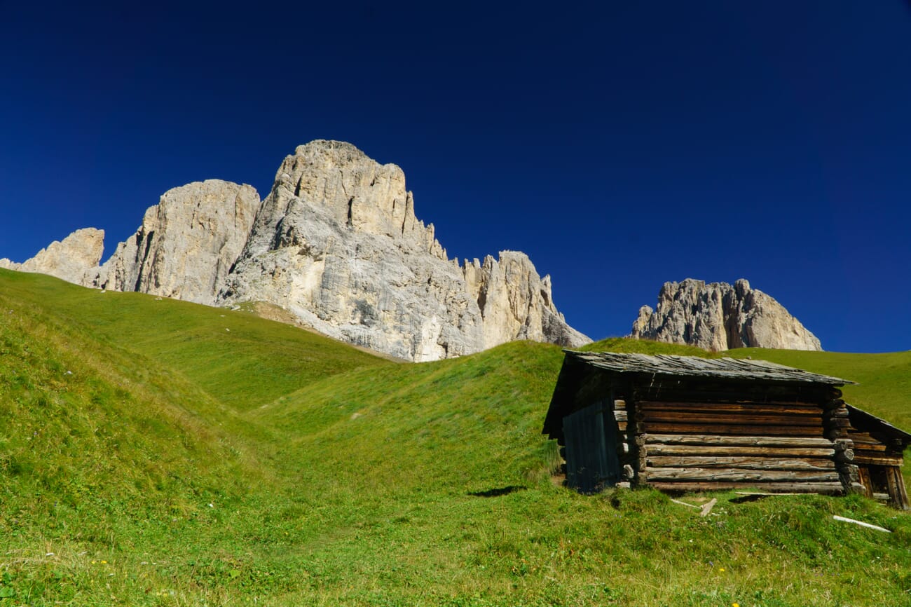 Wooden cabin on green grass with blue sky and sunlit mountains in the background.