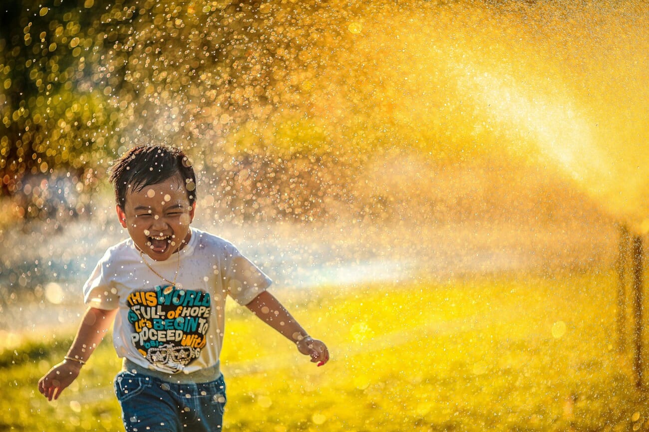 A young boy running joyfully through a spray of sprinkler water at golden hour, his smile sharp in focus while the background is a wash of warm gold and green.