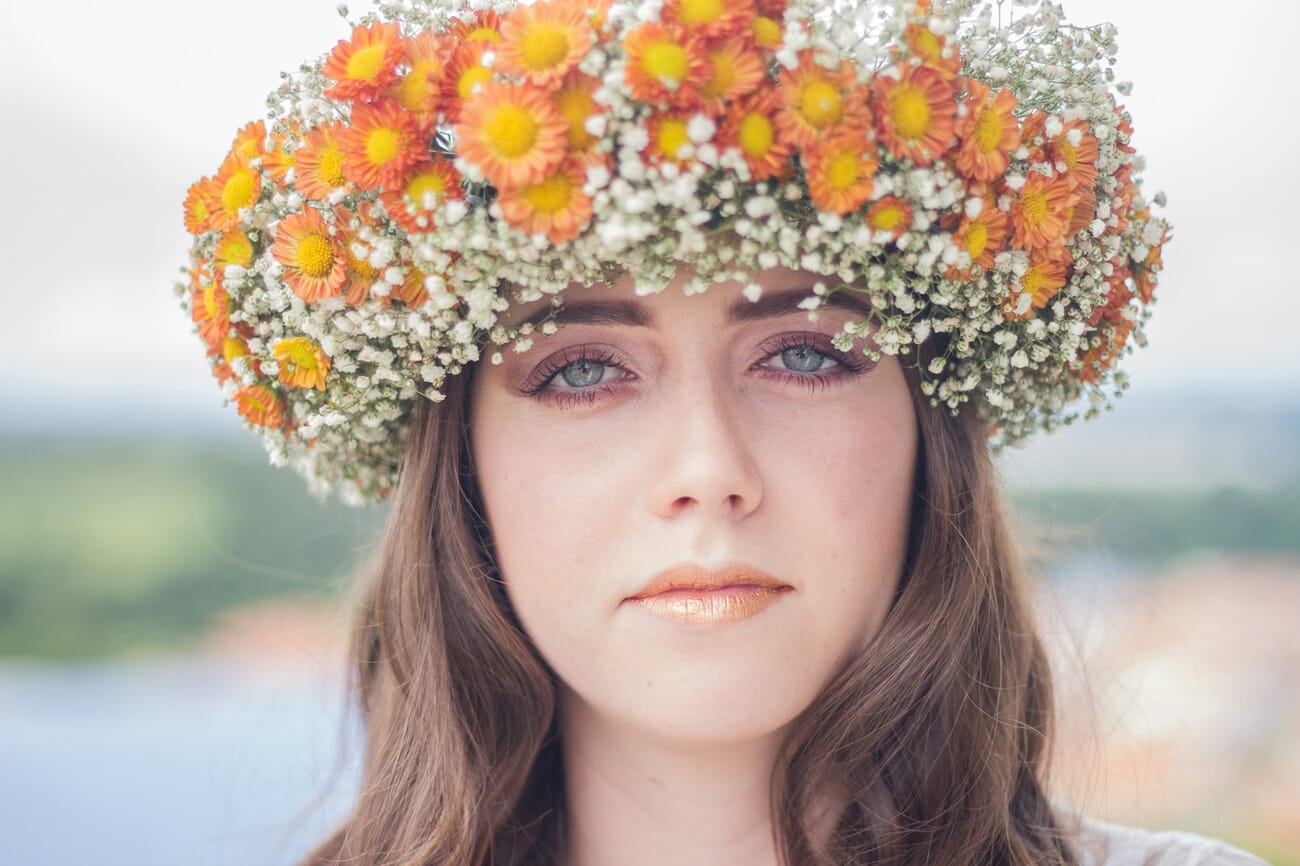 Close-up portrait of a woman with blue-green eyes wearing an orange marigold and baby's breath flower crown, soft green and blue background.