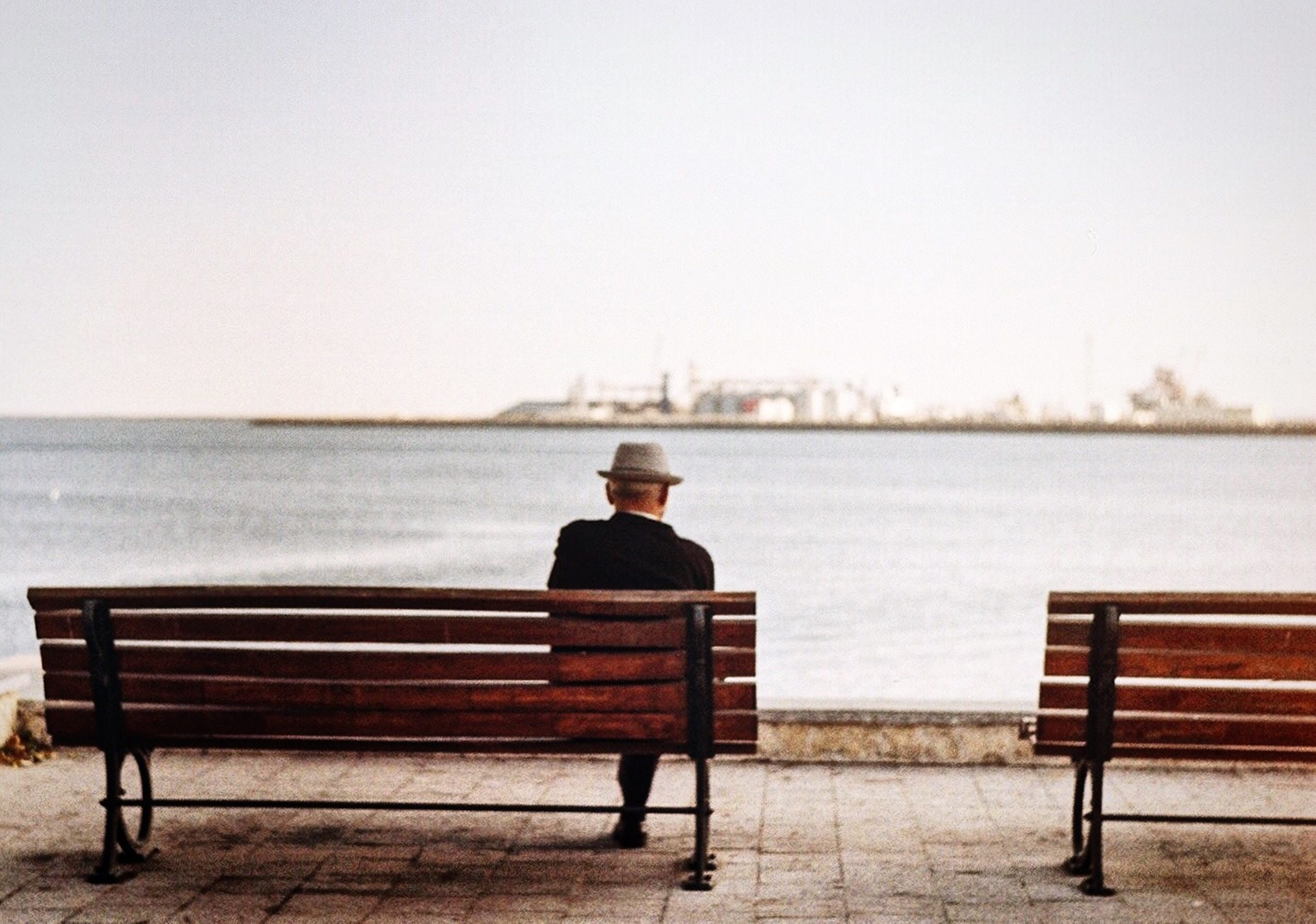 Man sitting on a bench by the water looking out at an industrial scene.