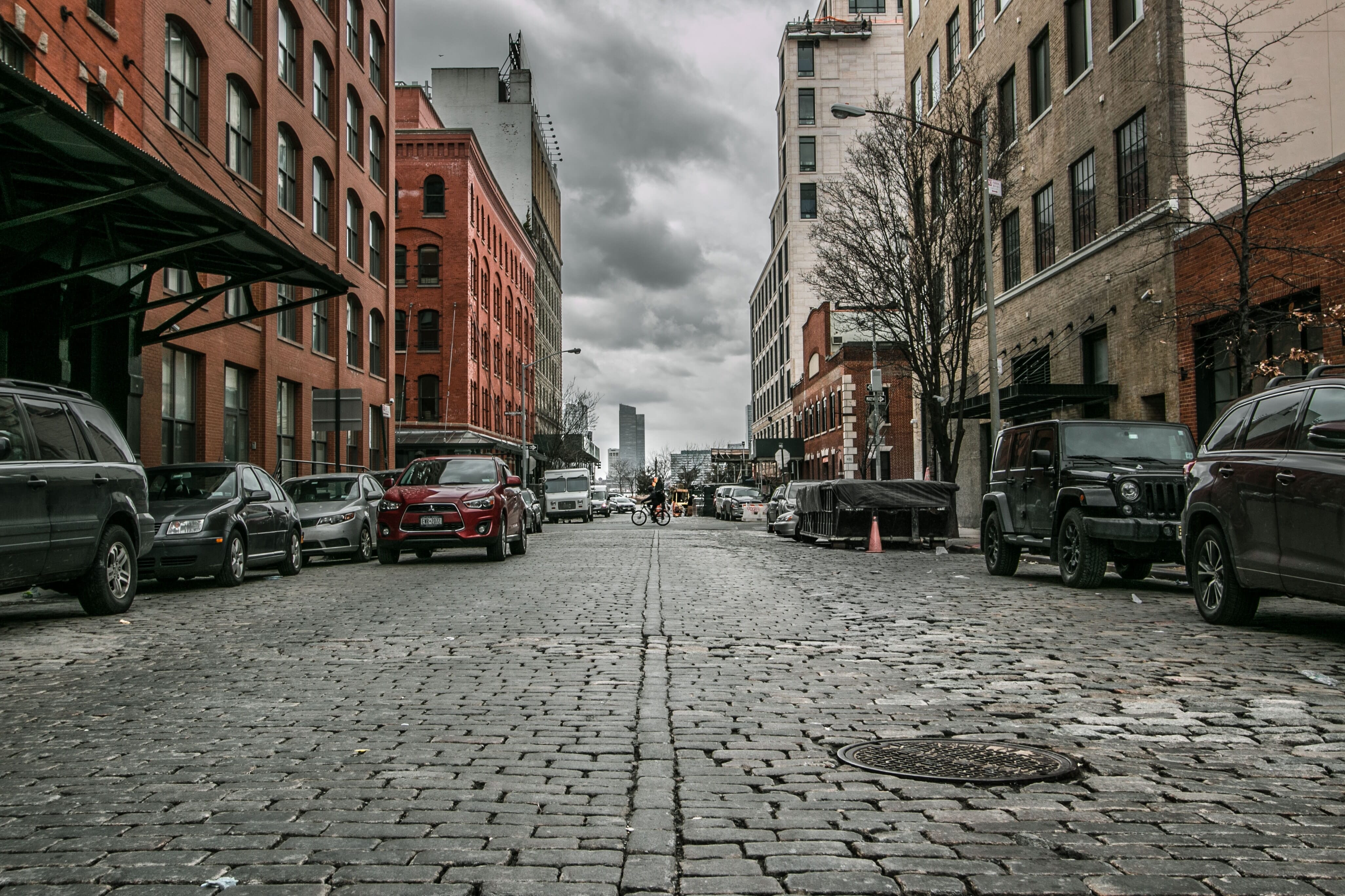 A street with cobblestones and lined with cars underneath stormy clouds.