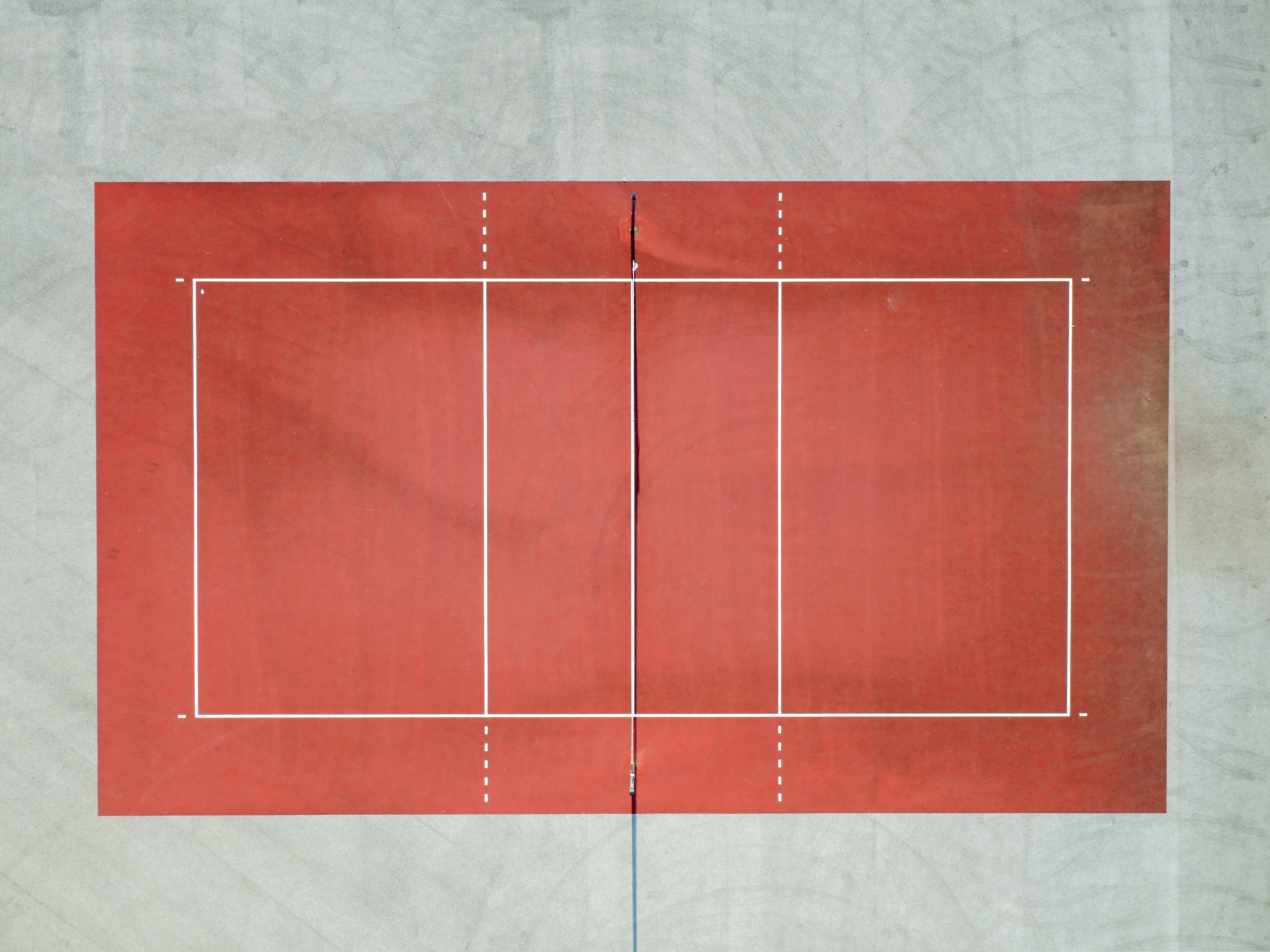 A red sports court viewed from above with a net in the middle and white surroundings.