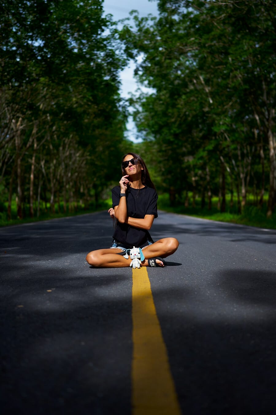 Woman sitting cross-legged on a tree-lined road in a relaxed criss-cross pose with one hand near her chin