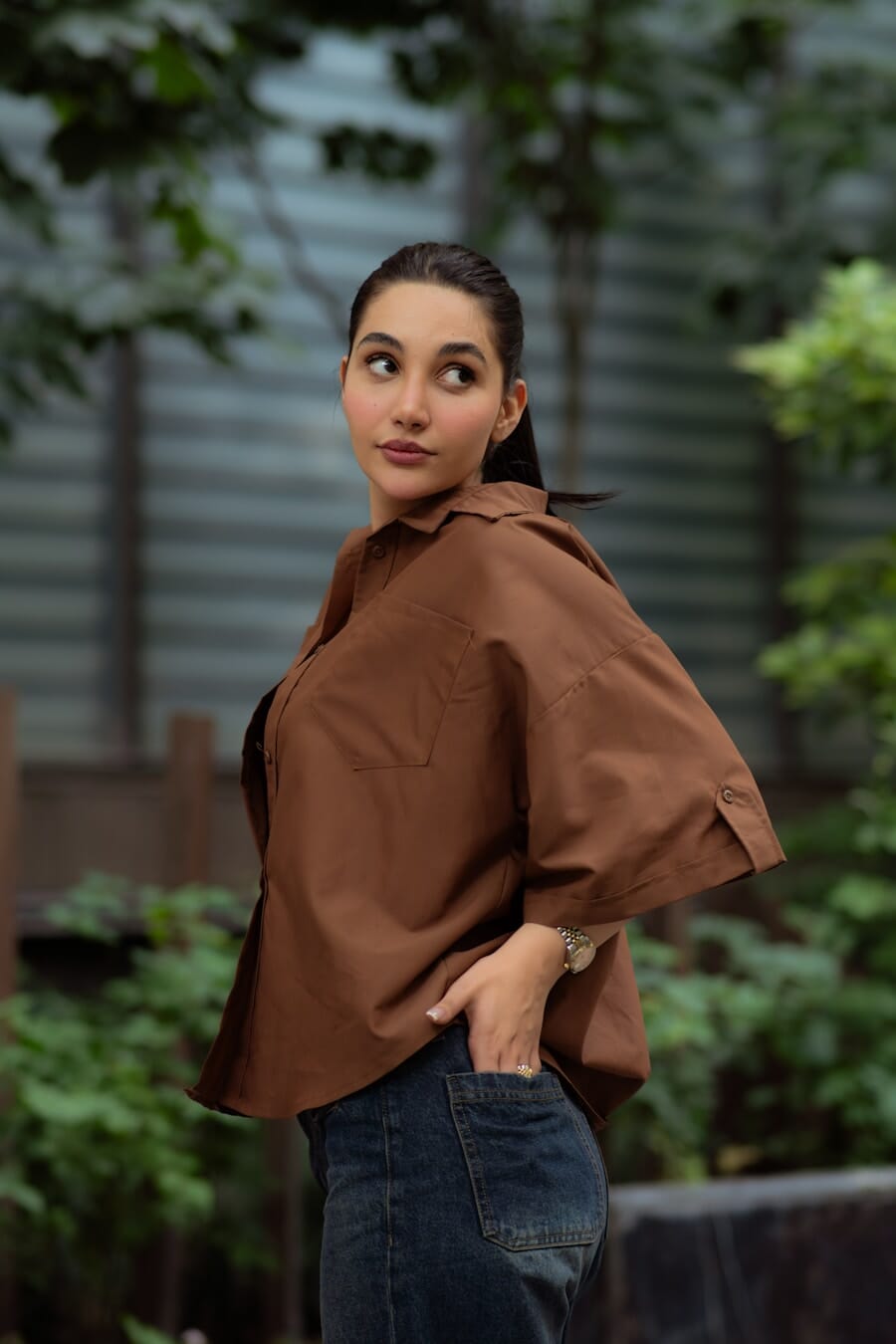 Woman in a brown shirt and jeans standing casually with hands in pockets