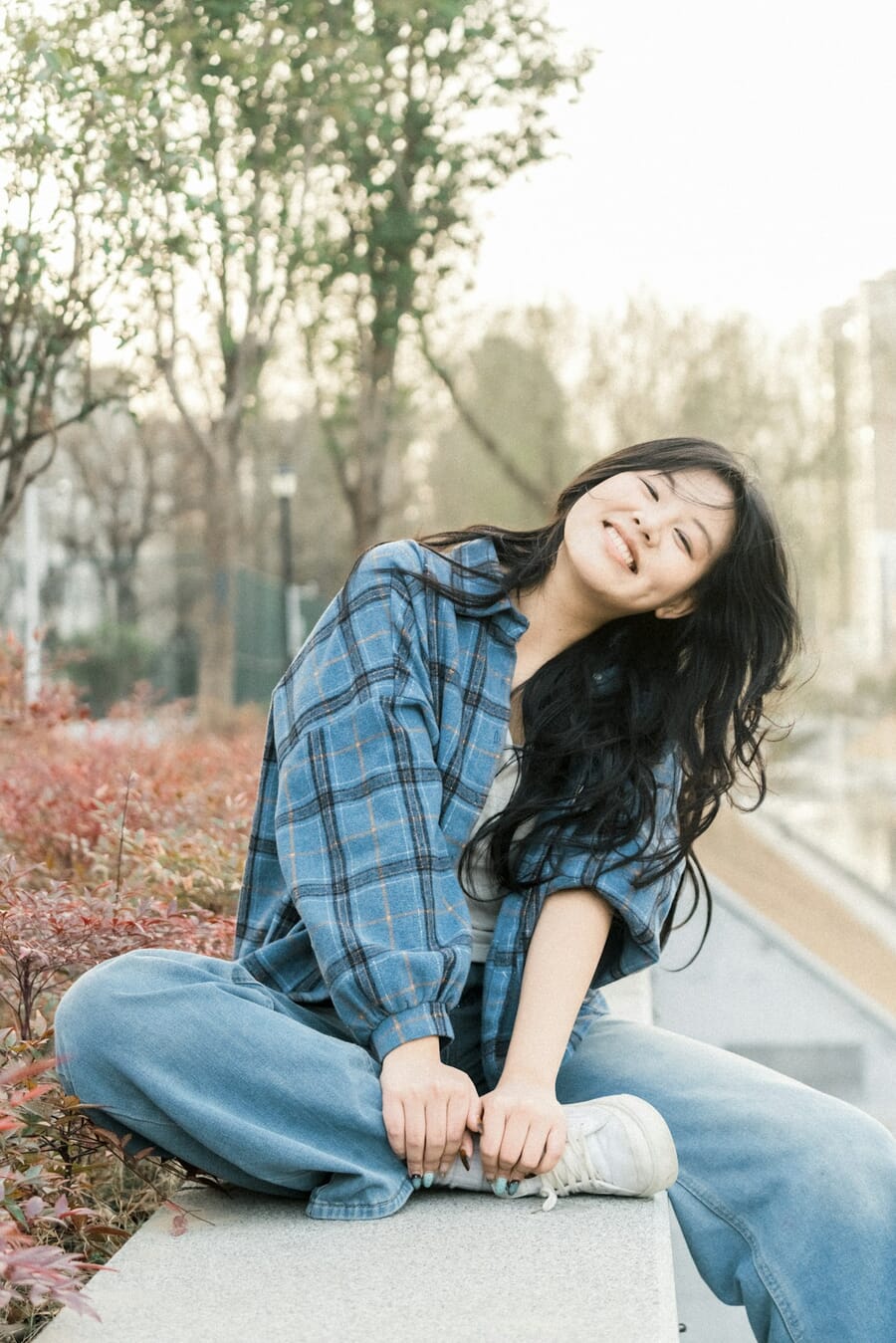 Young woman in a plaid shirt smiling warmly outdoors with a natural forward lean toward the camera