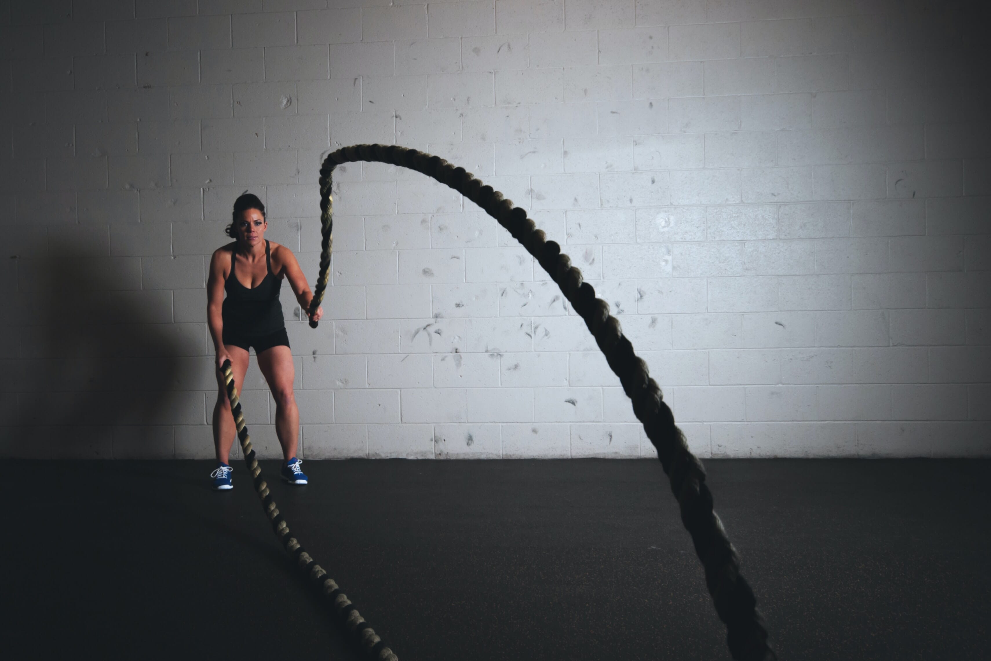 A woman dressed in black athletic clothes trains with ropes in front of a white wall.