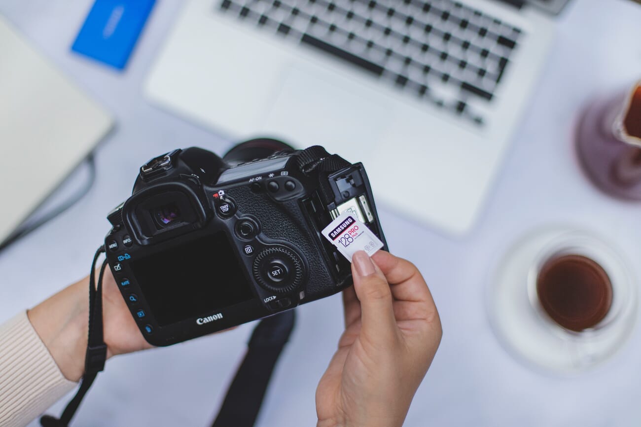 Hands removing a 128 GB SD memory card from a black Canon DSLR camera with a laptop in the background