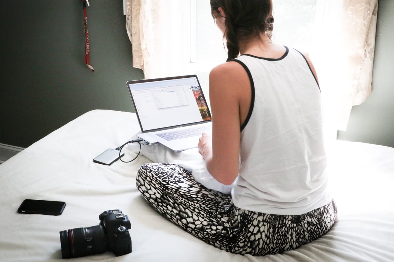A woman sitting on a bed with a laptop, external hard drive, and camera doing a photo backup session