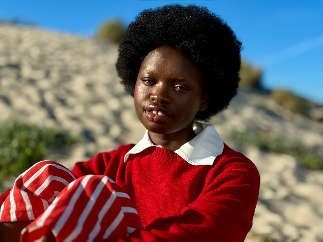 iPhone 17e sample portrait of a woman with an afro in a red and white top against a sandy background, shot at 2x telephoto