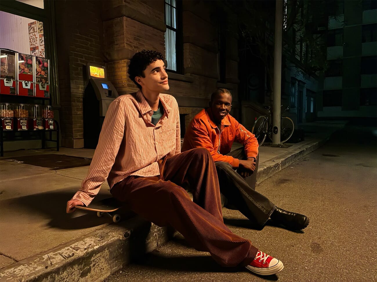 iPhone 17e sample photo of two people sitting on a stoop under a neon convenience store sign at dusk, showing Night mode