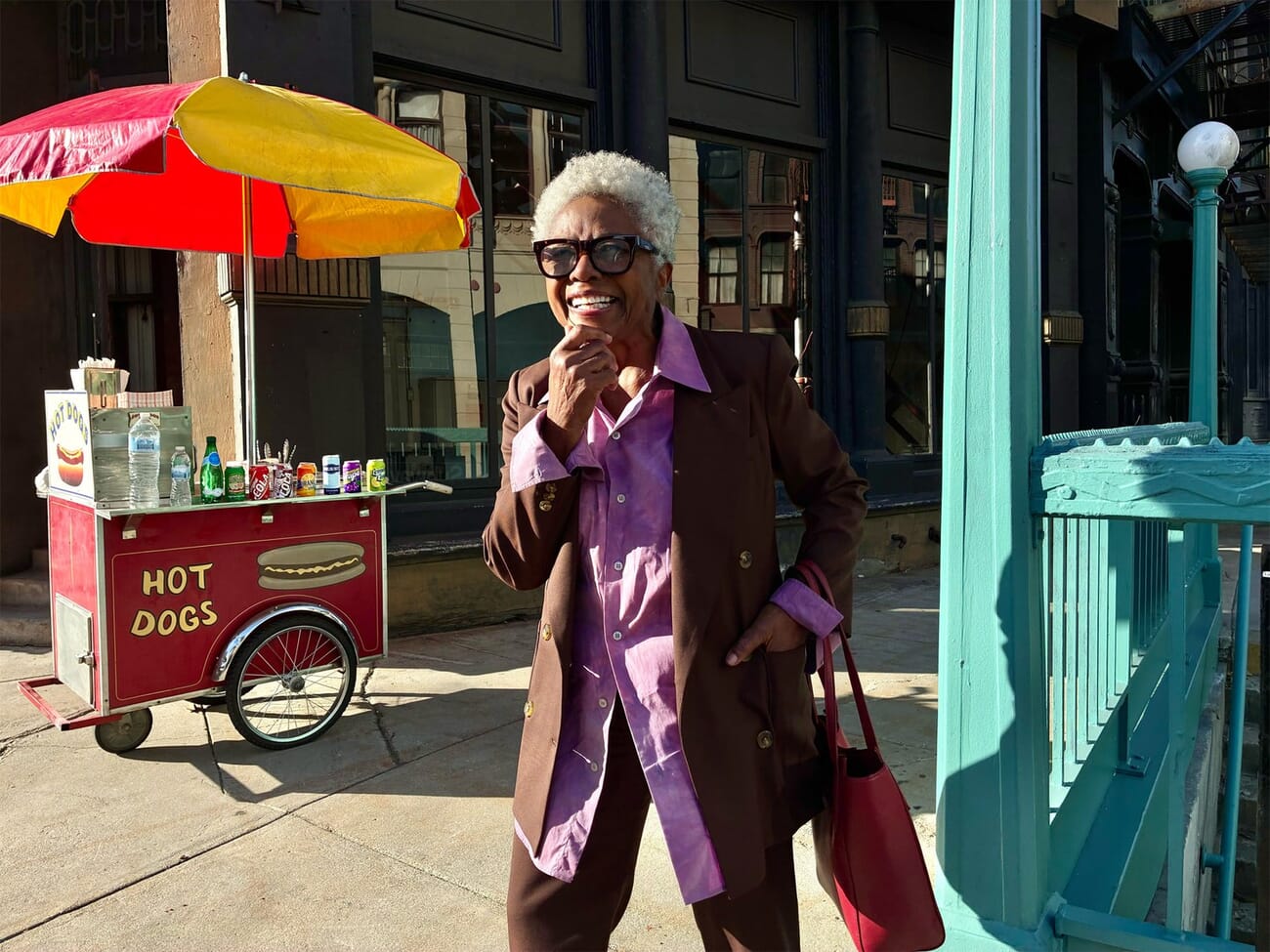 iPhone 17e sample photo of a street vendor in a purple velvet blazer holding shopping bags in front of a hot dog cart