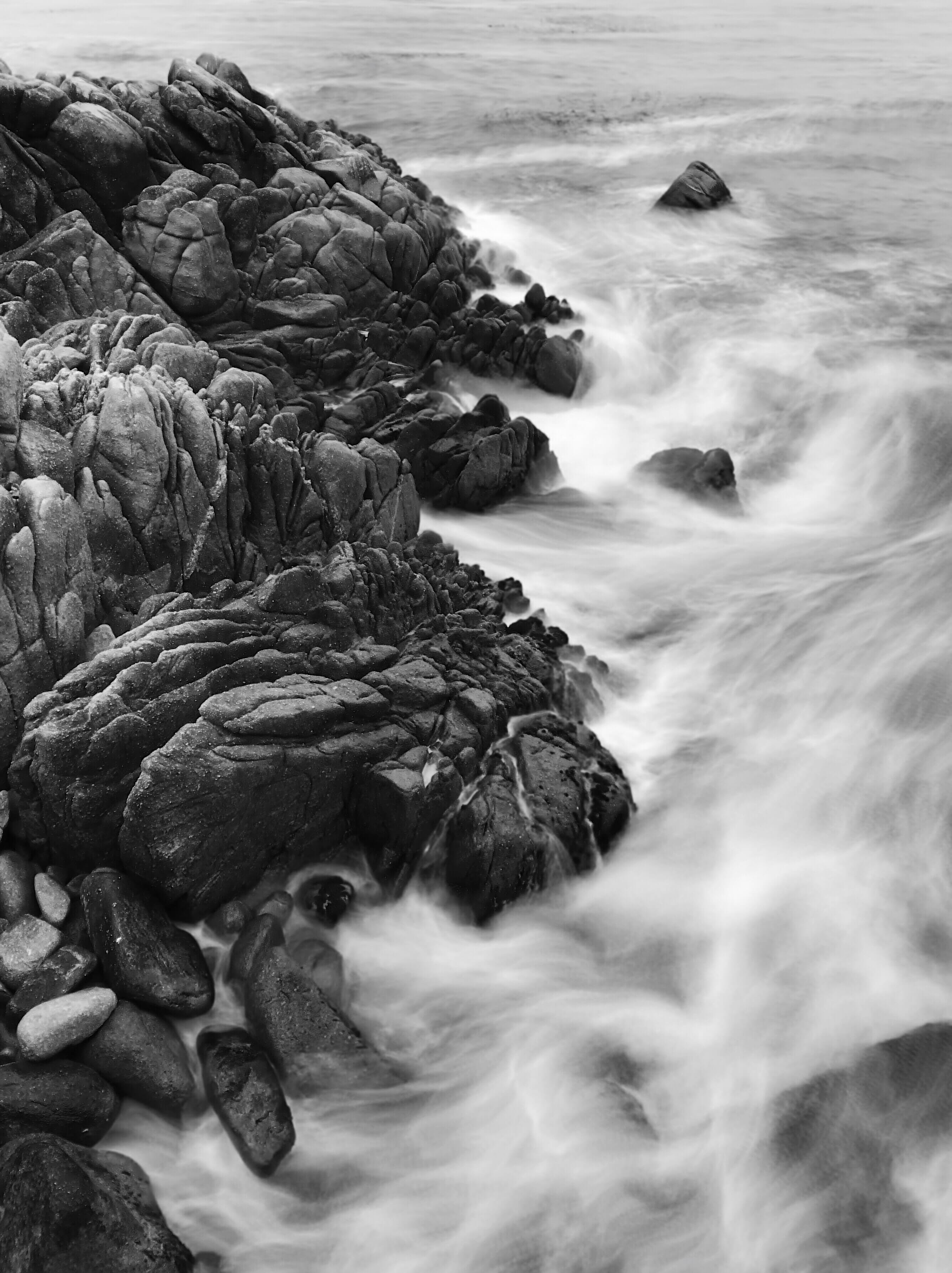 A moody black-and-white seascape with rugged rocks on the left and swirls of water on the right.
