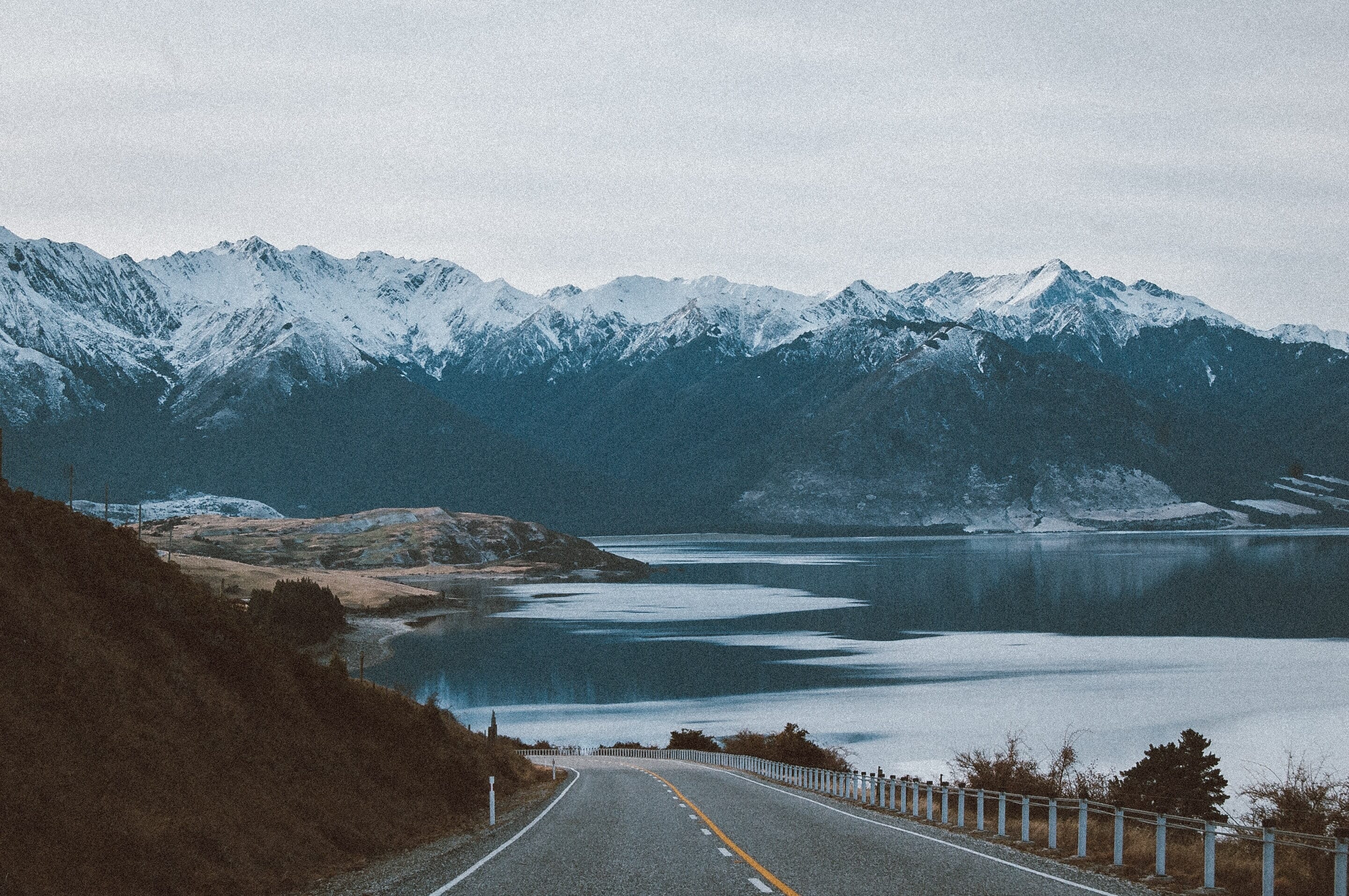 A road stretches off toward a partially frozen lake and snowy mountains beneath a gray sky.