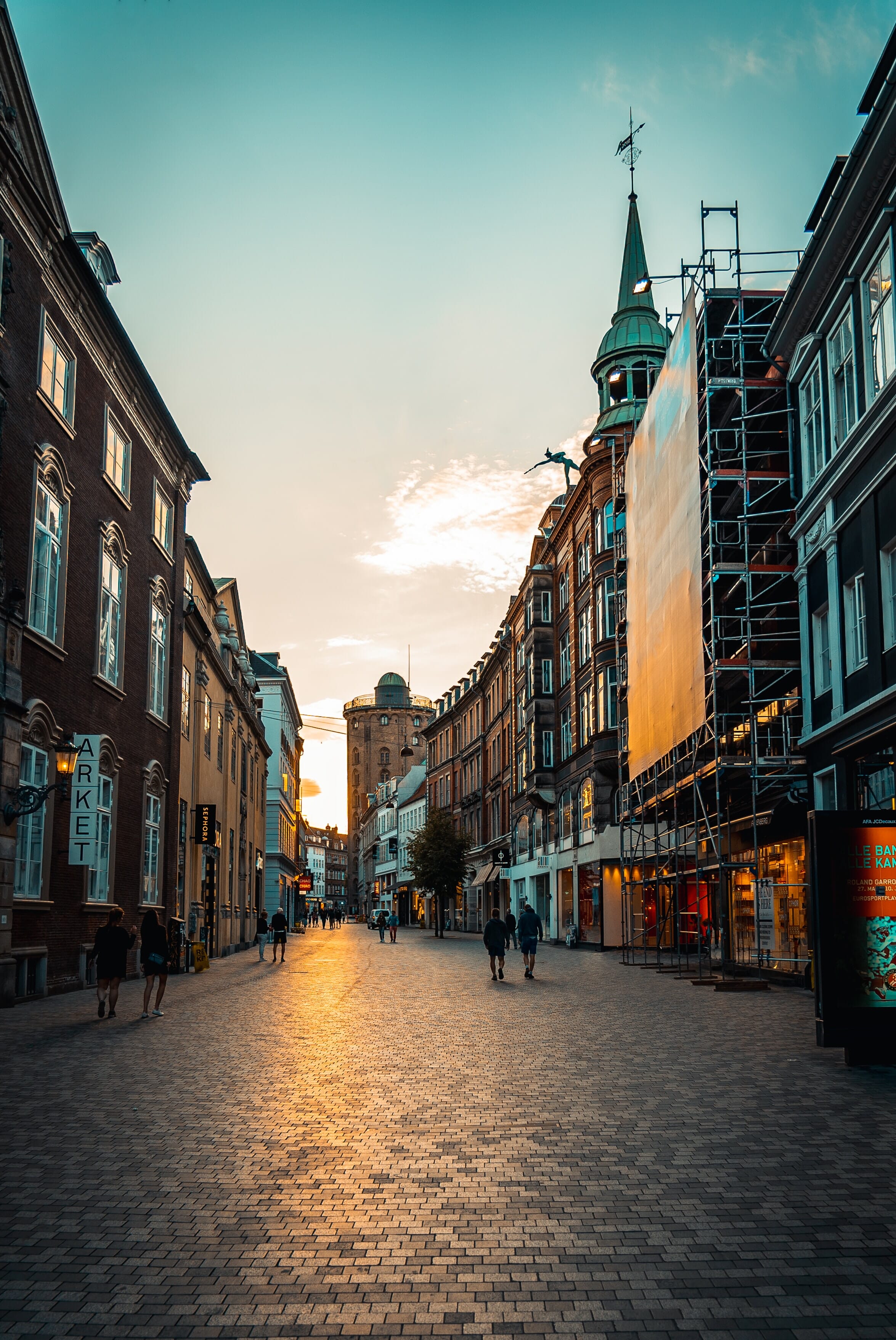 A narrow city street at sunset with people walking by shops.