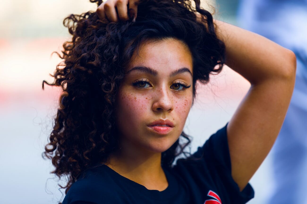 A natural-light portrait of a young woman with dark curly hair, her face sharply in focus against a softly dissolved outdoor background, shot at f/2.8 · 1/200 · ISO 125