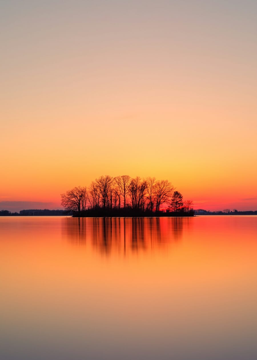 Dark silhouettes of trees on a small island against a fiery orange sunset sky with reflection in water