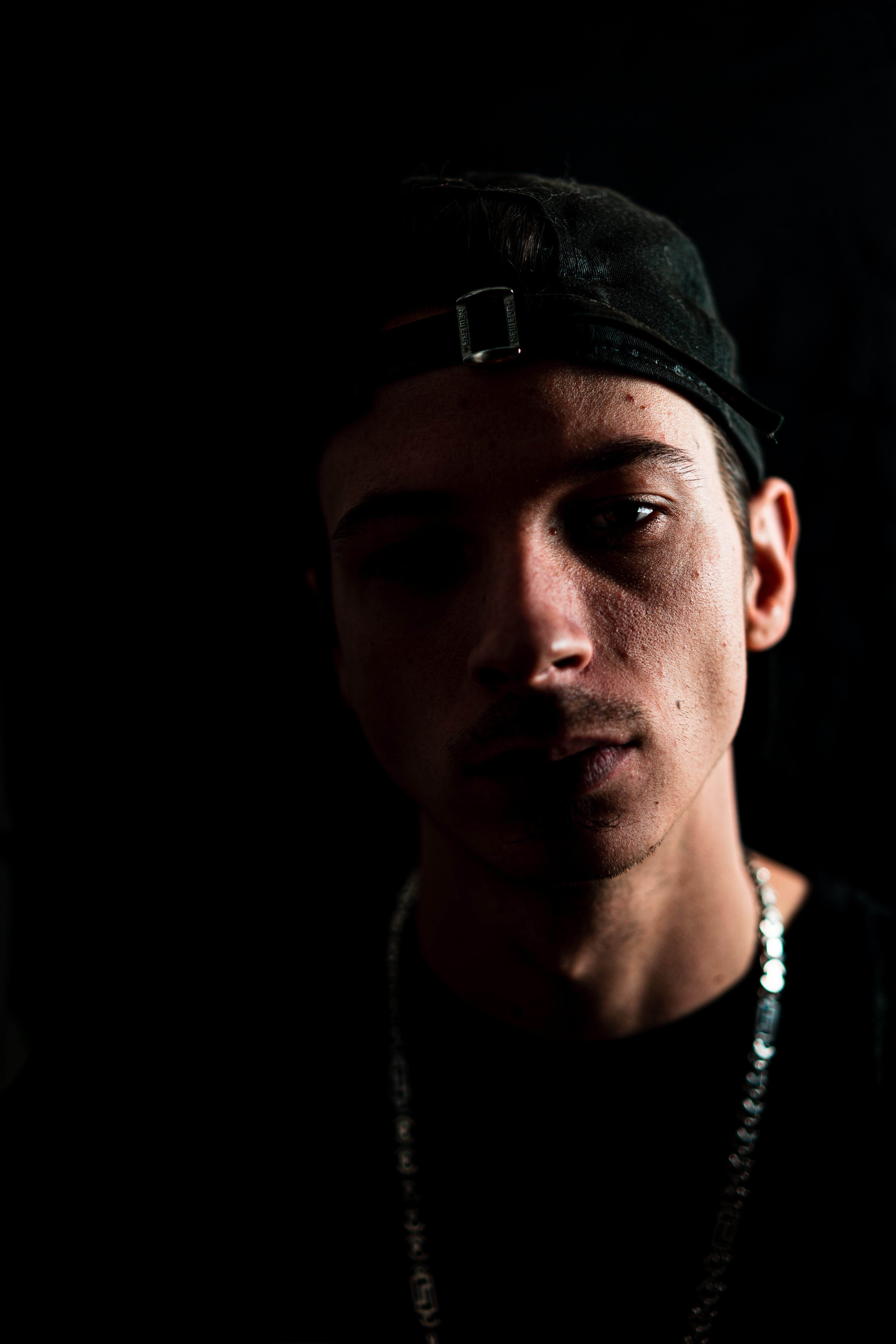 A man with a backward cap and a short mustache partially lit against a black background.
