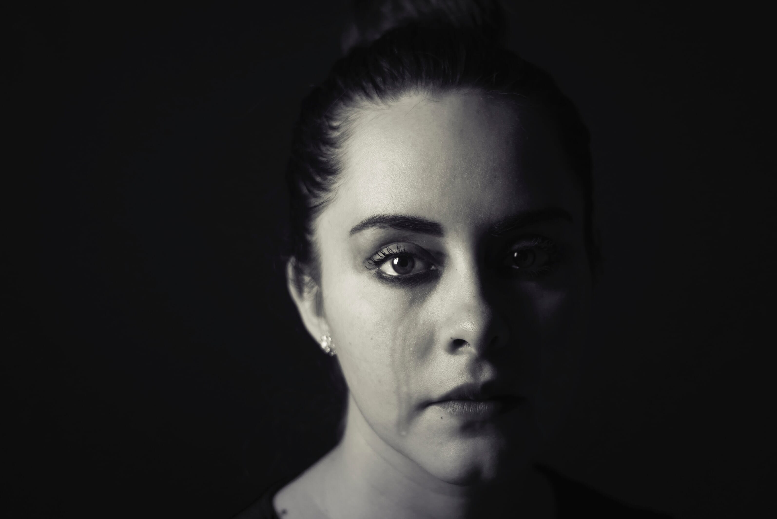 A dark-haired woman on a black background with a tear running down her cheek in black and white.