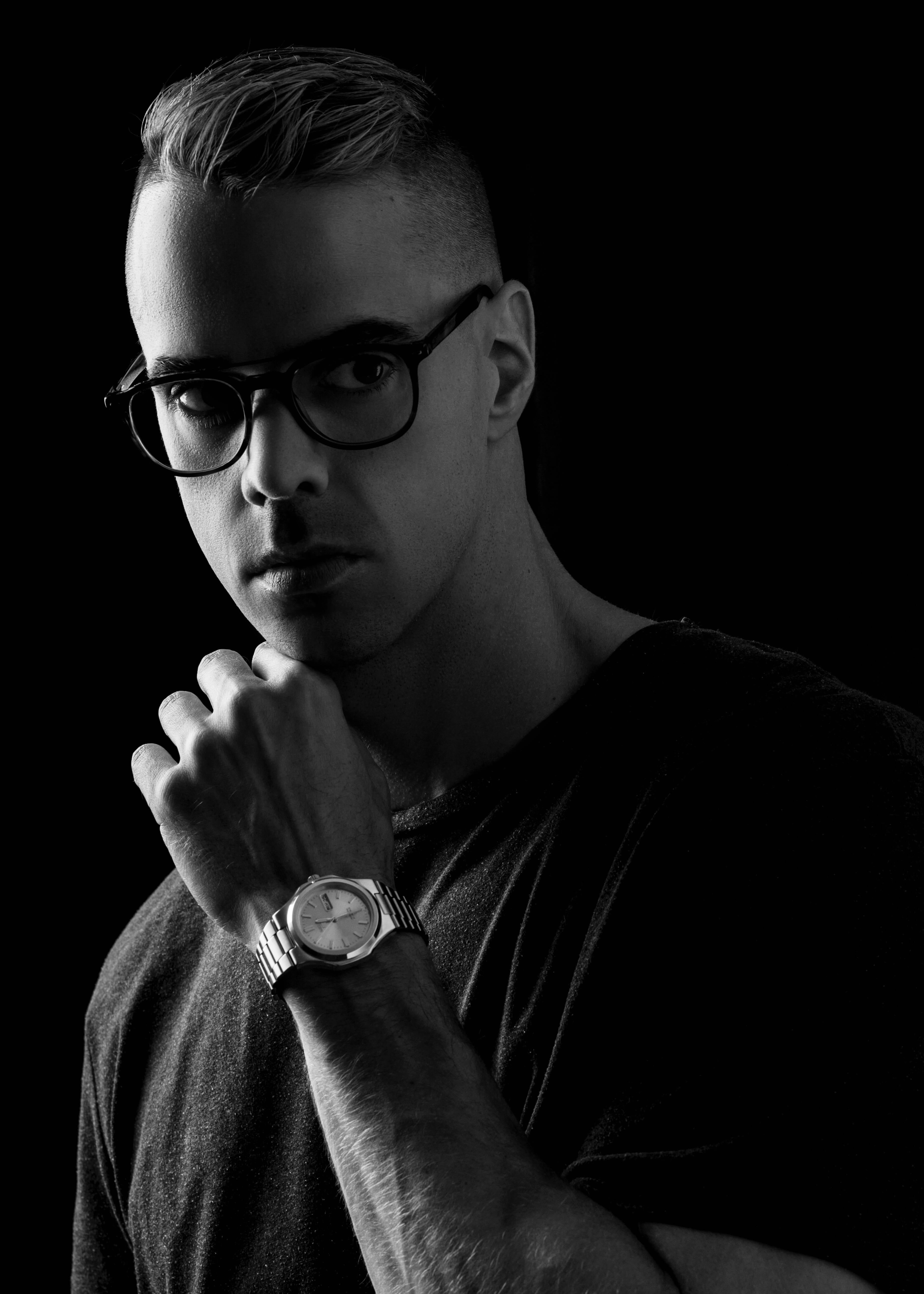 A man with short hair and glasses with a hand under his chin on a black background.
