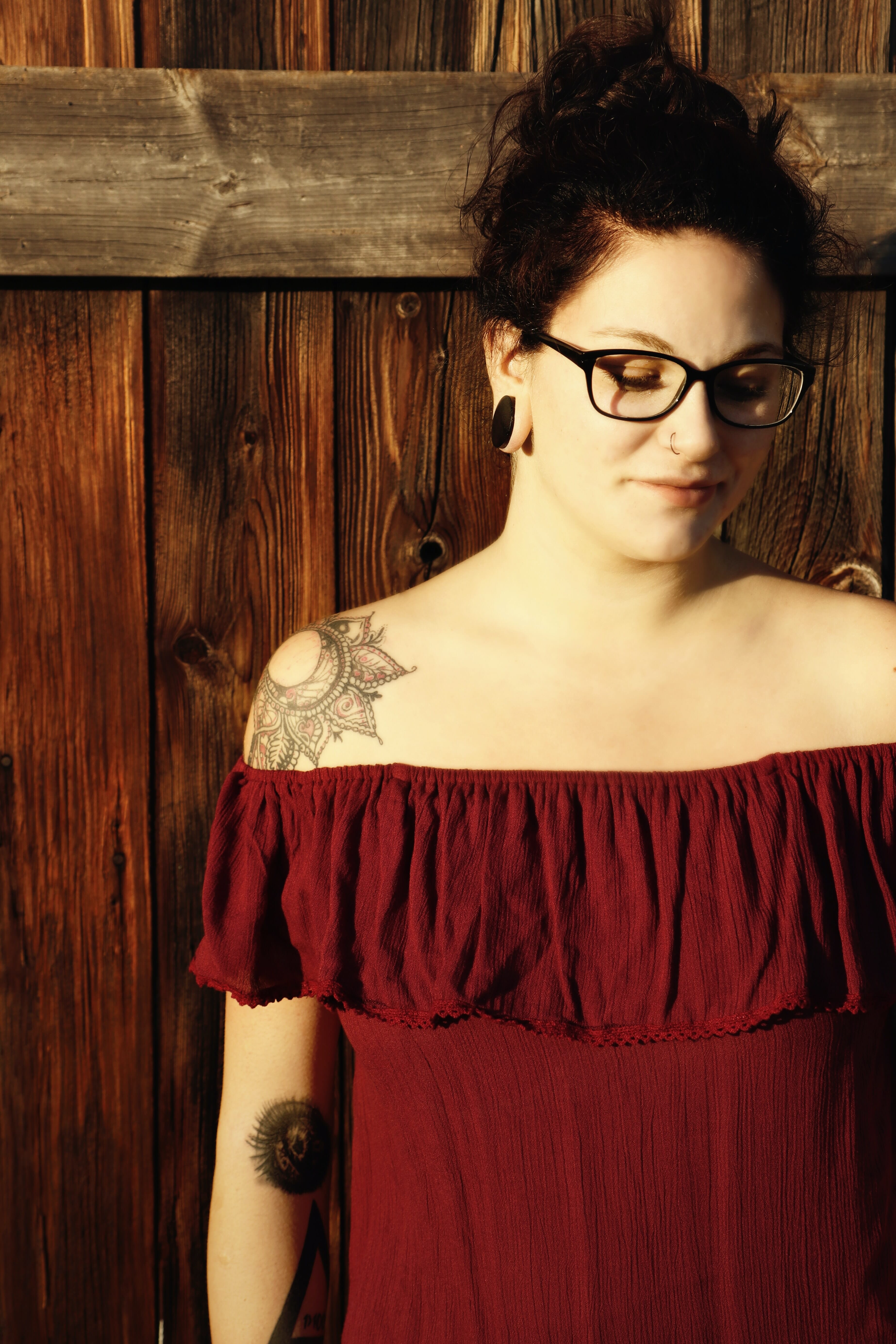A dark-haired woman with a nose ring, black glasses, and large black earrings in a red dress against a rustic wooden background.