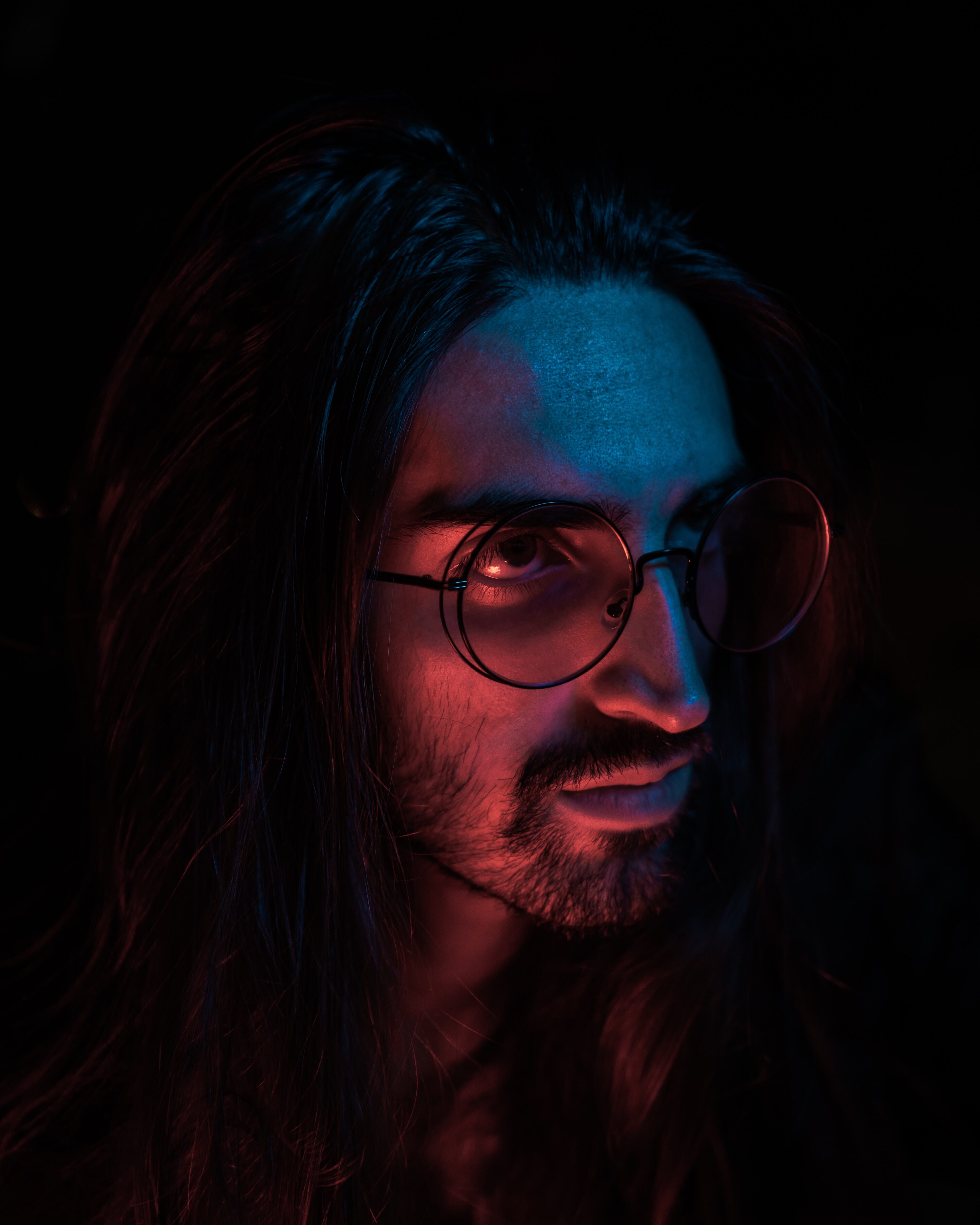 A man with long hair, a mustache, and a beard wearing round glasses lit by blue and red light and surrounded by darkness.