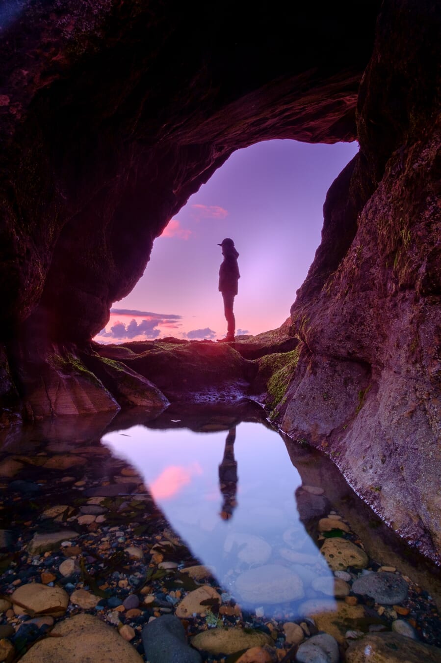 A person at the mouth of a cave at sunset reflected in a rocky tide pool