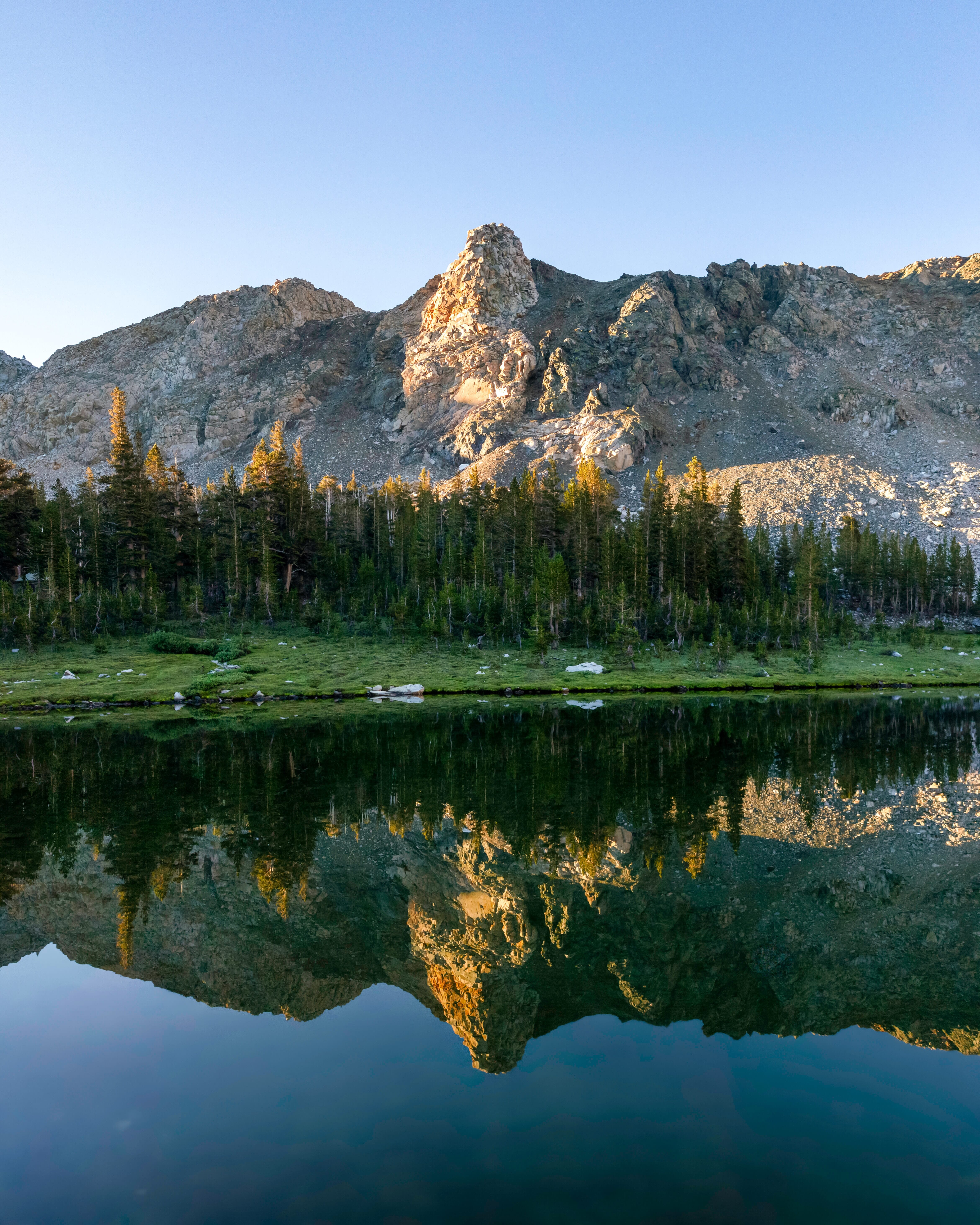 A tranquil lake reflects a small forest and mountain in the morning sunlight.