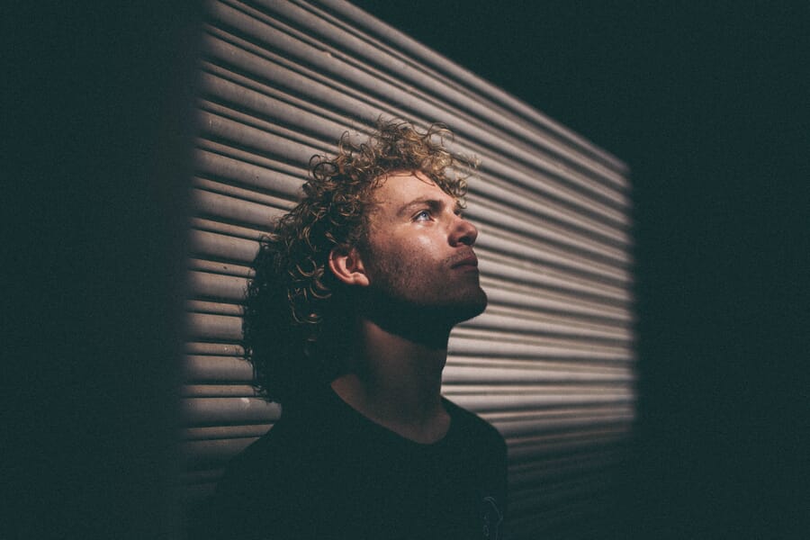 Curly haired man looking up while leaning back against a metal surface surrounded by darkness