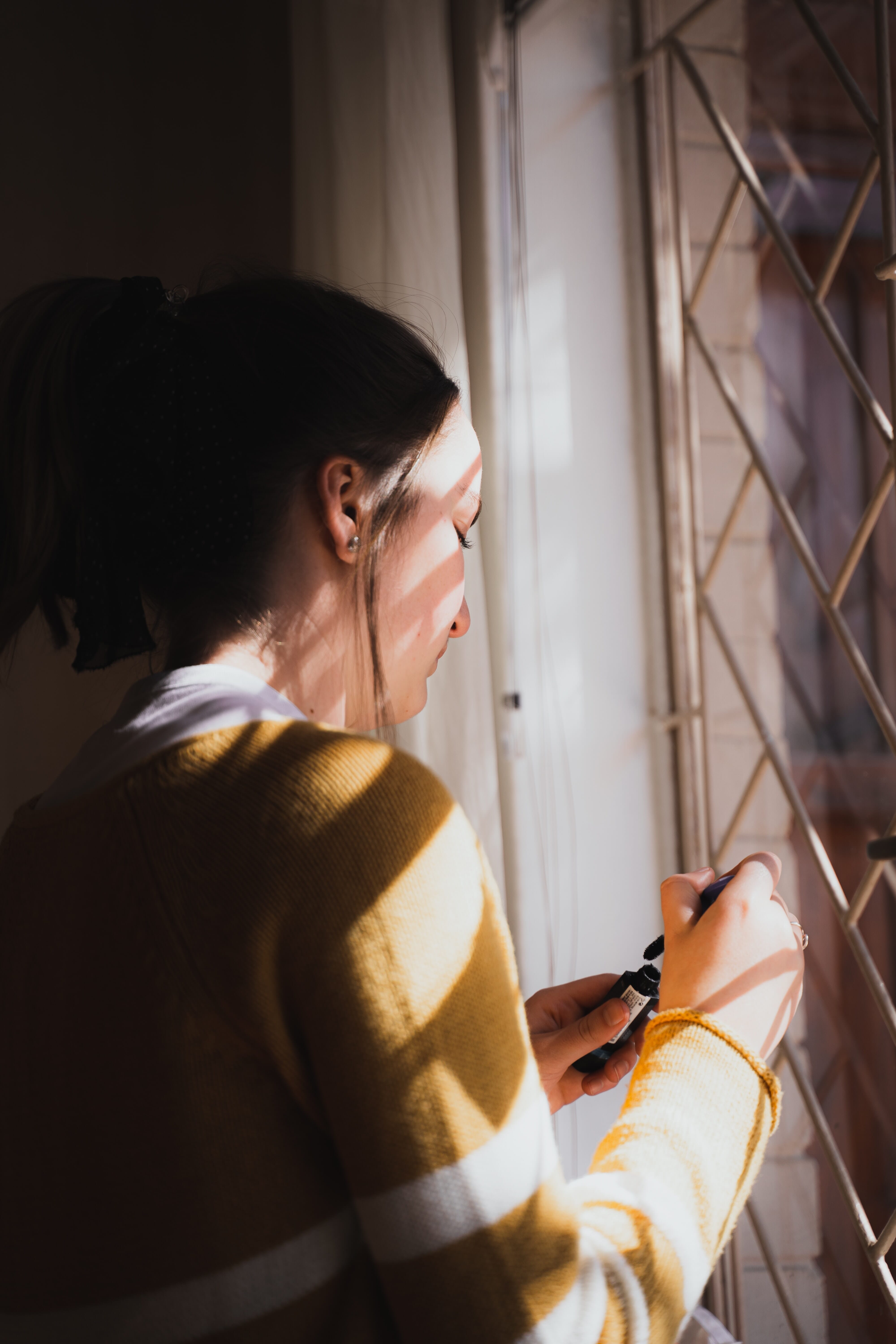 Dark-haired woman in a yellow top standing in front of a window holding a small black bottle.
