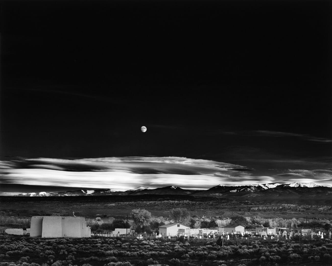 Ansel Adams original 1941 black-and-white photograph Moonrise, Hernandez, New Mexico: a full moon over an adobe village and cemetery with mountains and a luminous cloud band behind.