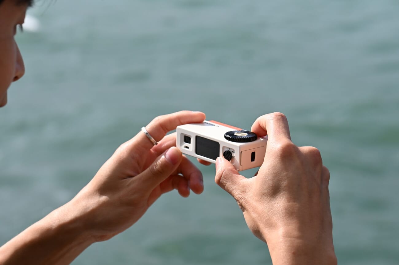 A person framing a shot two-handed with the Yashica Y16 in orange, by the water