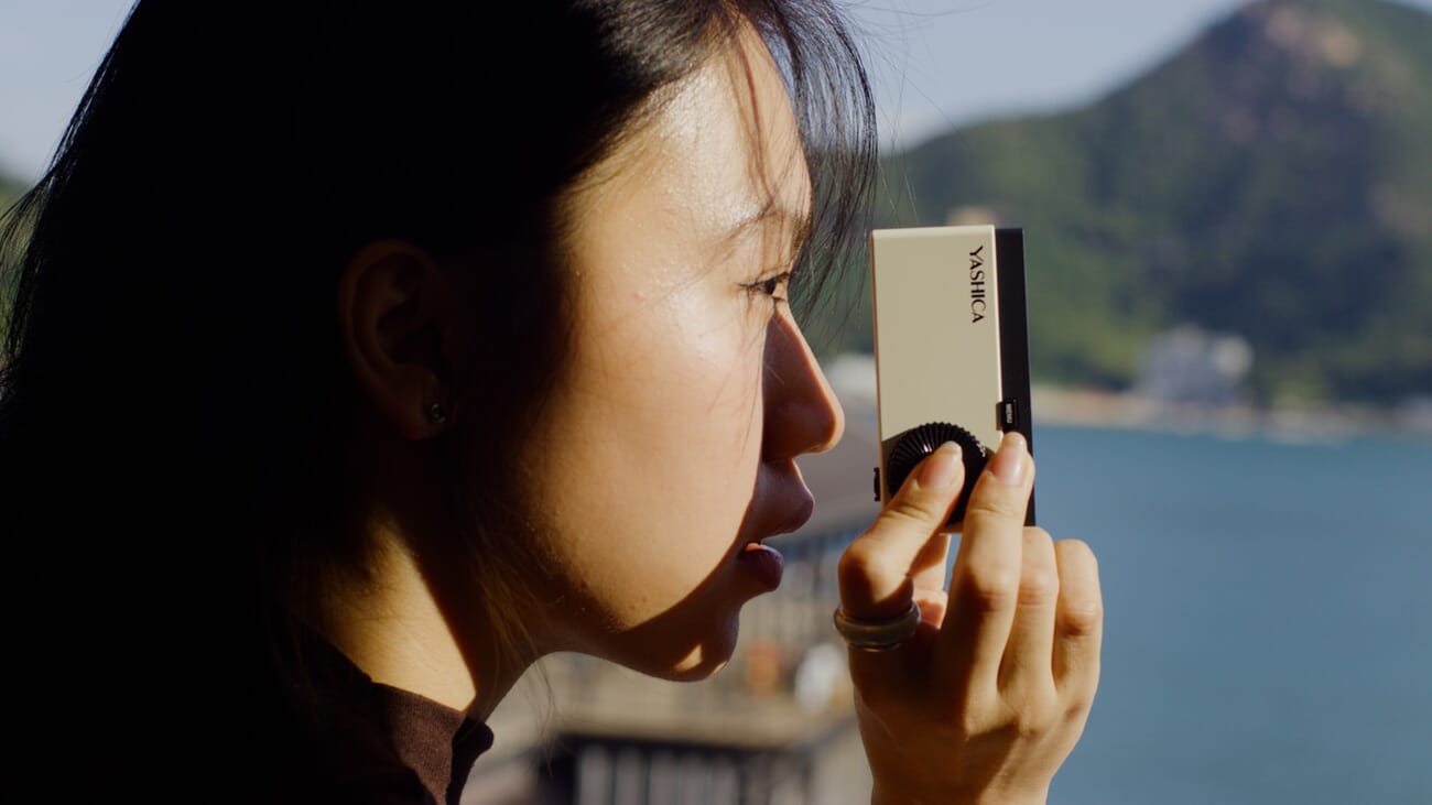 A person holding the Yashica Y16 up to their eye to use its optical viewfinder, with a coastal backdrop