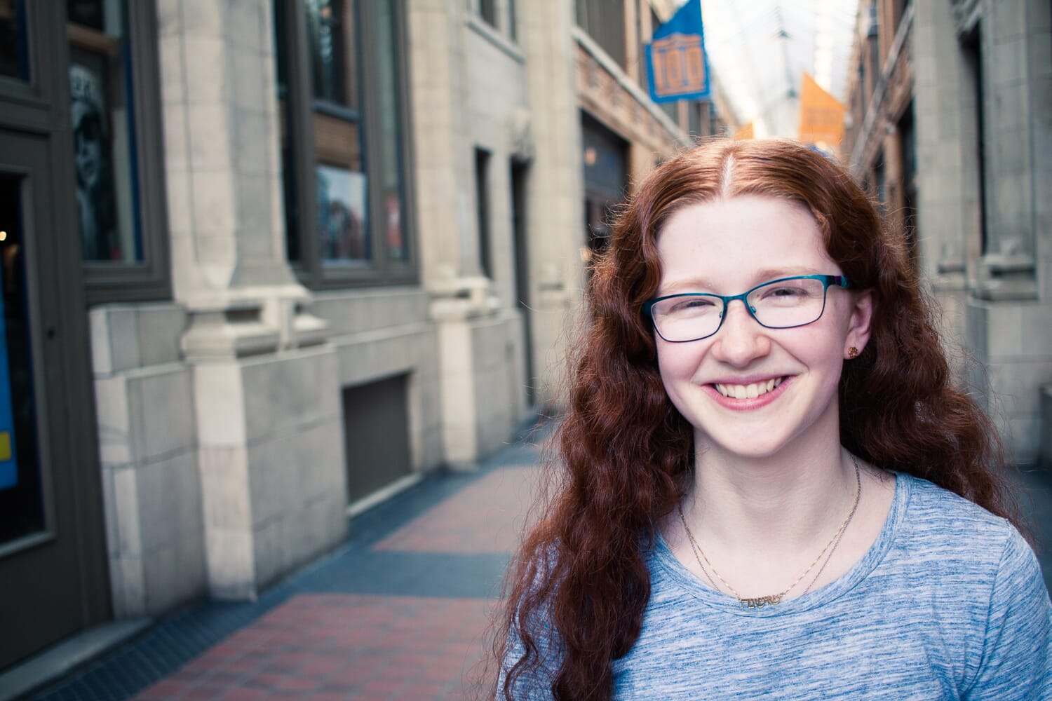 Girl with red hair and a blue top smiling with a corridor in the background.