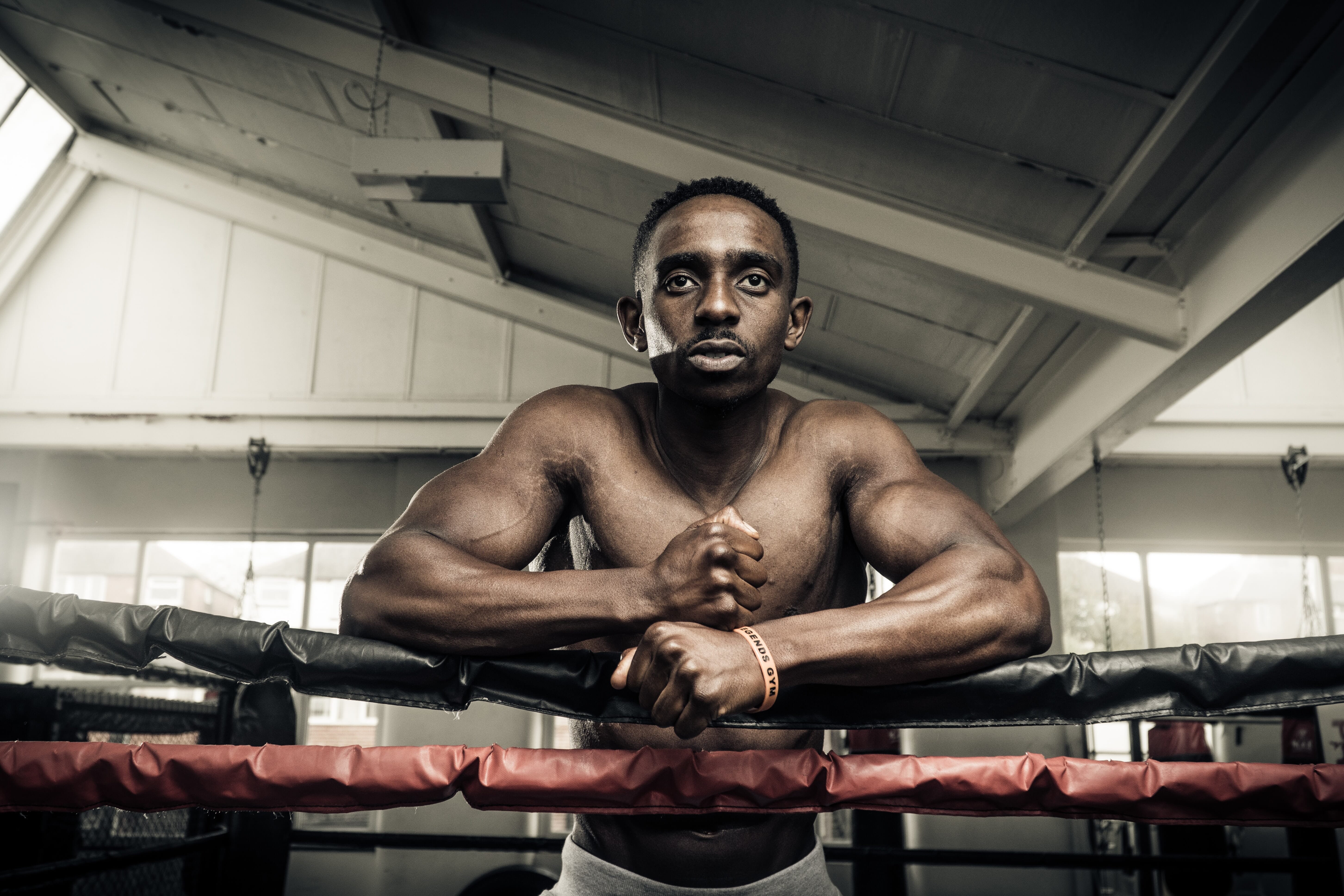 A focused boxer leans on a black and red ring, showcasing muscular arms. Natural light floods a gym, emphasizing determination and strength in the athletic.