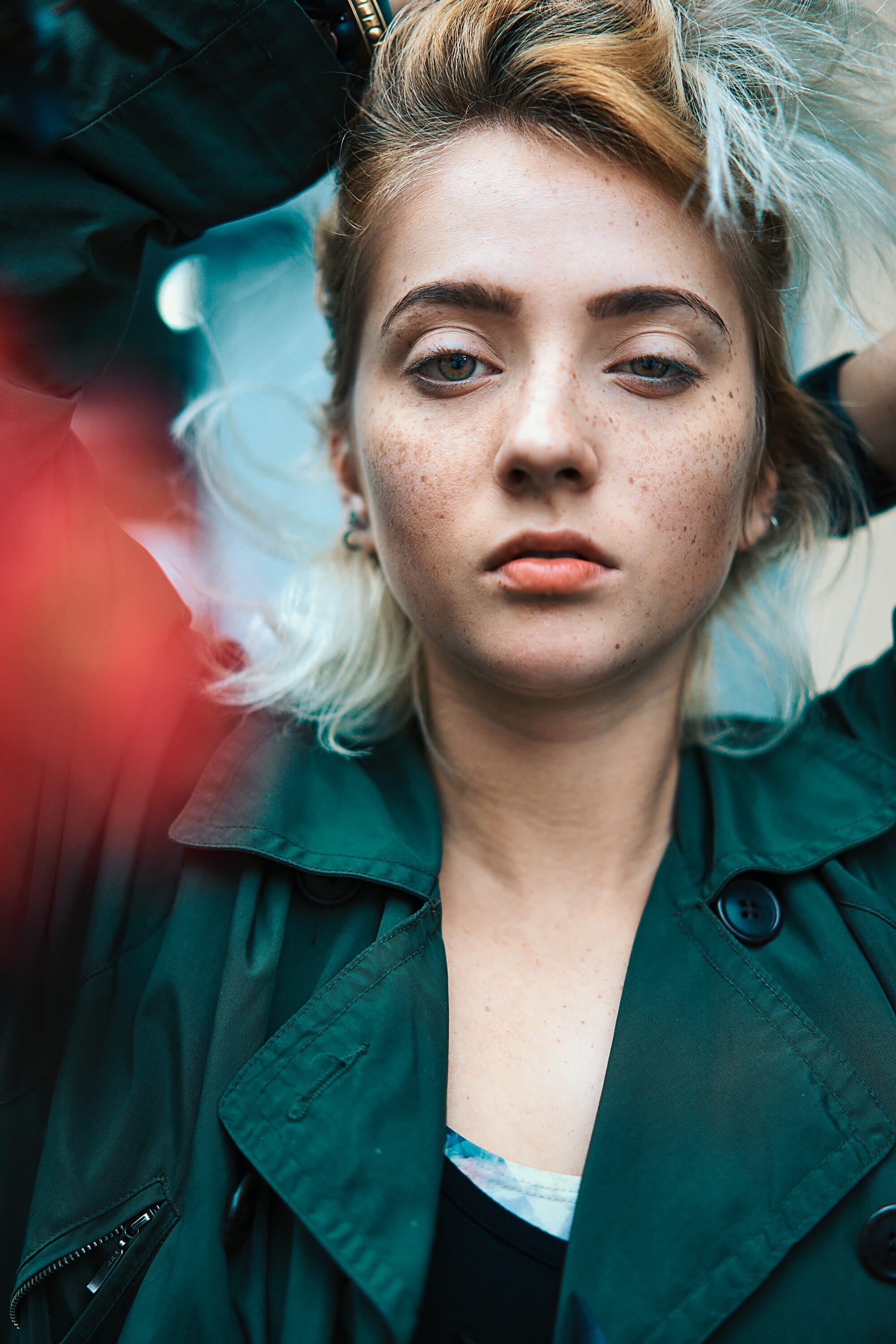 A close-up portrait of a model with striking blue eyes and freckles, dressed in a green trench coat. The image captures soft lighting and a blurred floral background, emphasizing her expressive.