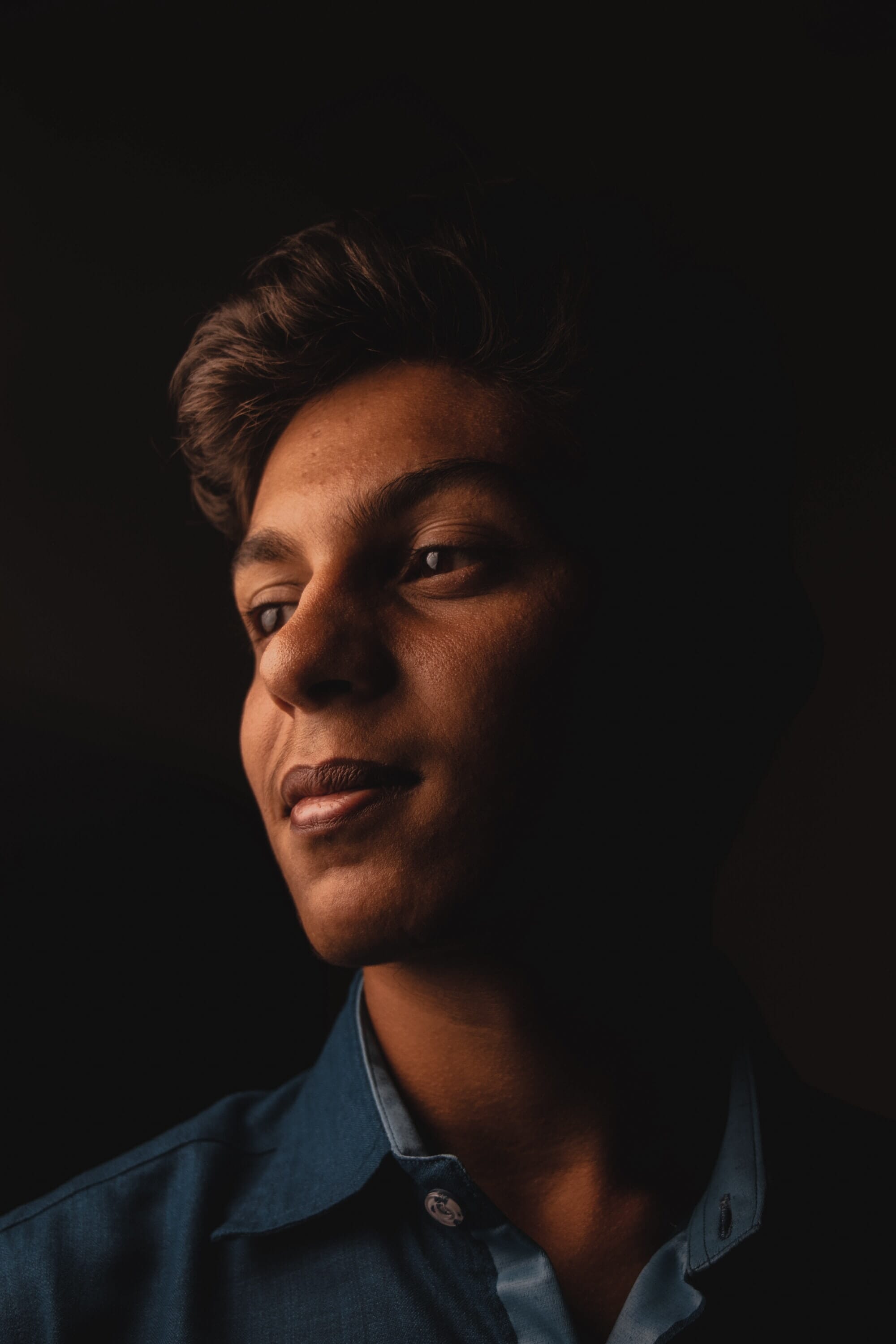 A close-up portrait of a young man in dramatic lighting, with a dark background highlighting his features. The soft glow accentuates his expression and textured shirt. Ideal for exploring external flash.