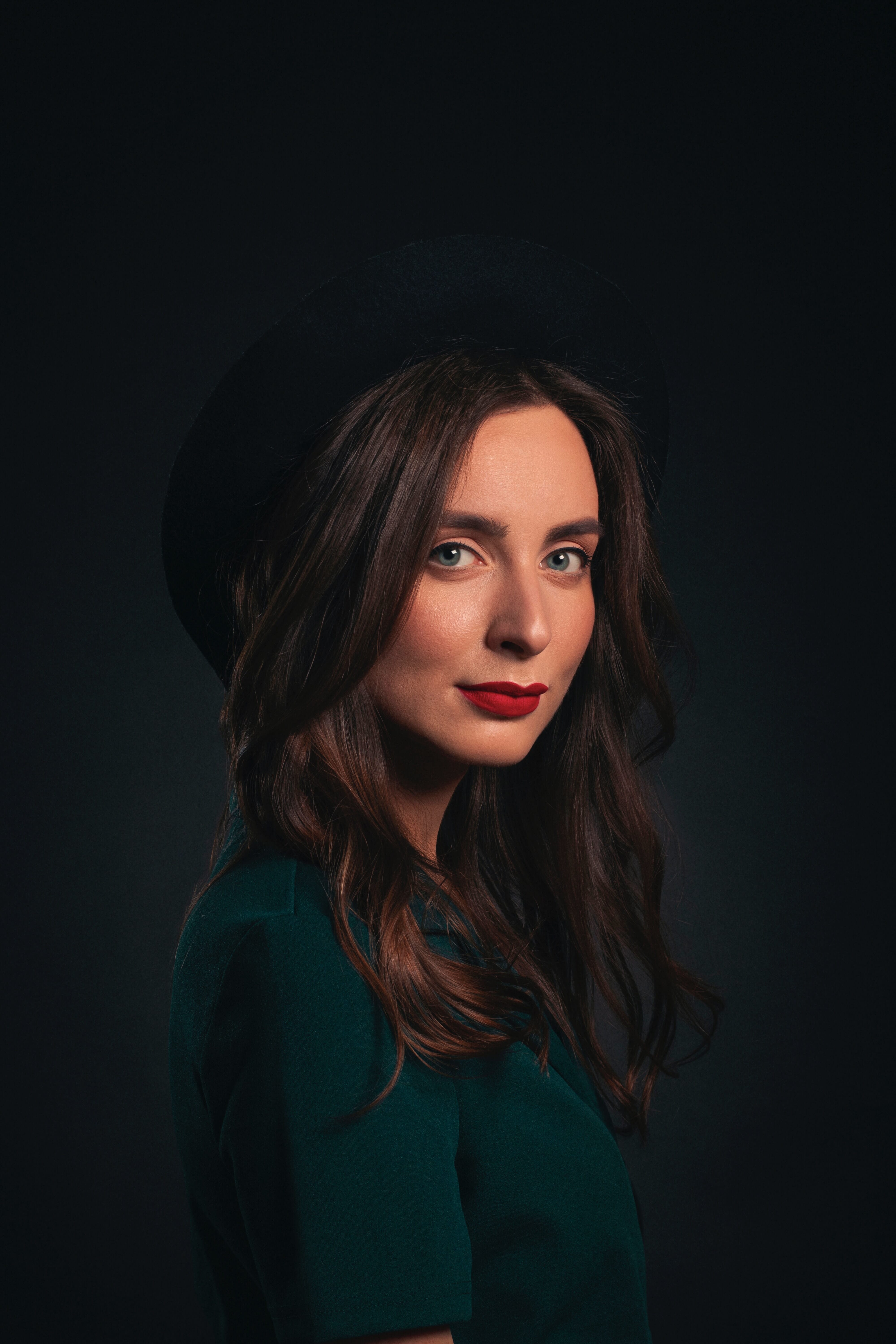 Elegant portrait of a woman in a dark setting, showcasing rich brown hair, striking red lips, and a stylish black hat, highlighting effective use of external flash for dramatic.