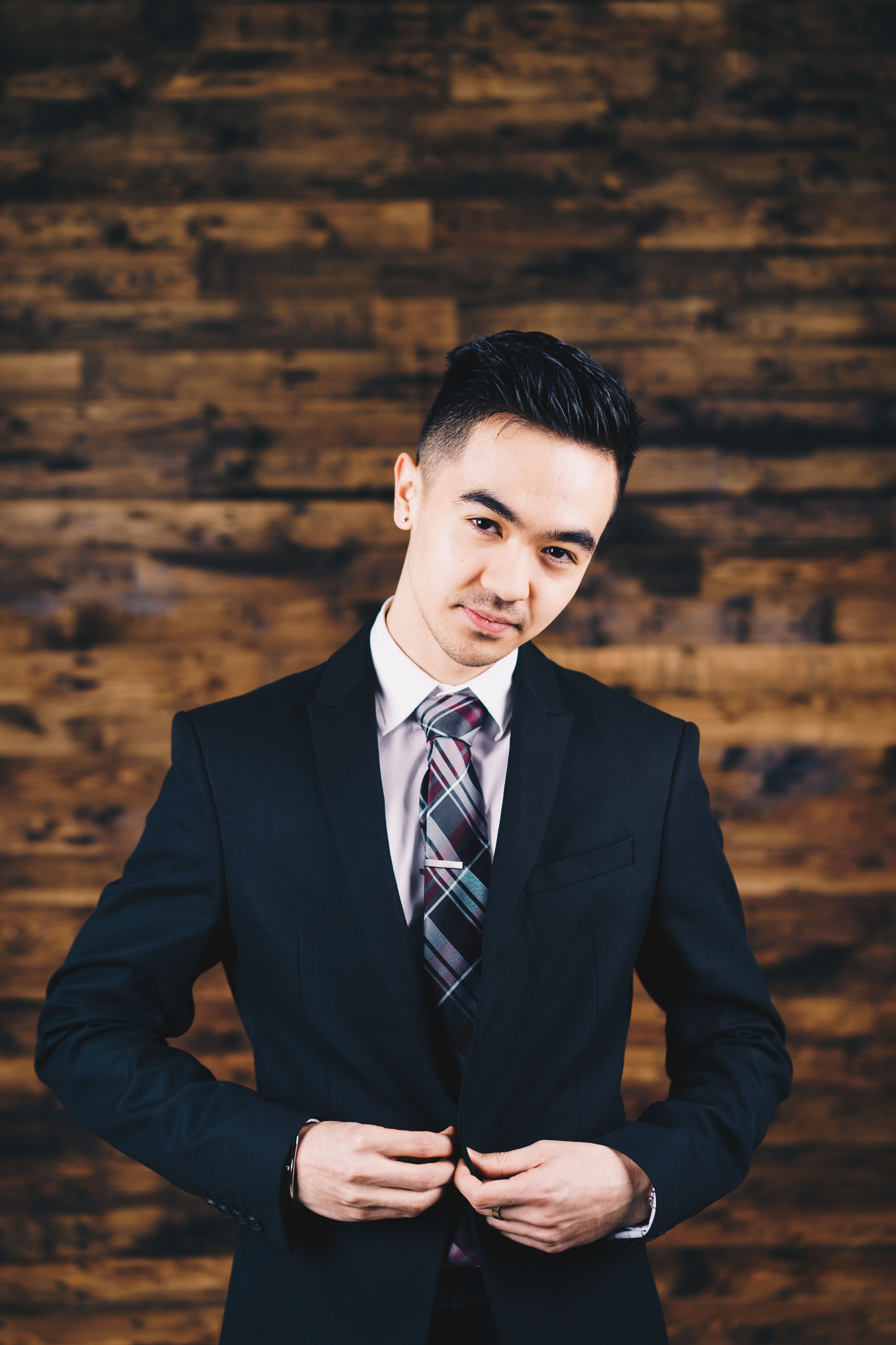 A man with short, styled hair buttoning his suit jacket while standing in front of a brown backdrop.