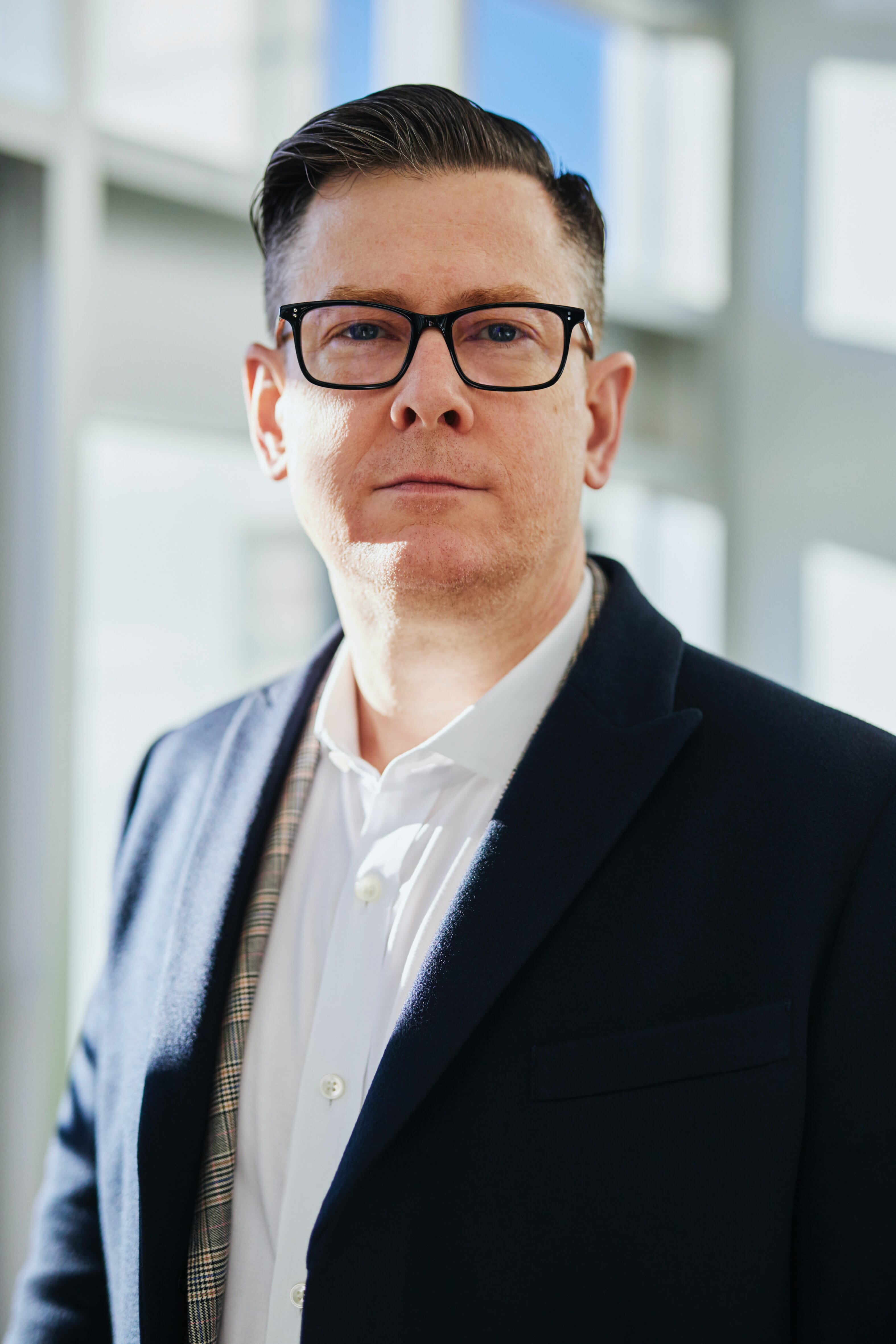 Headshot of a suit-wearing man with slicked-back hair and glasses looking at the camera with blurry windows in the background.