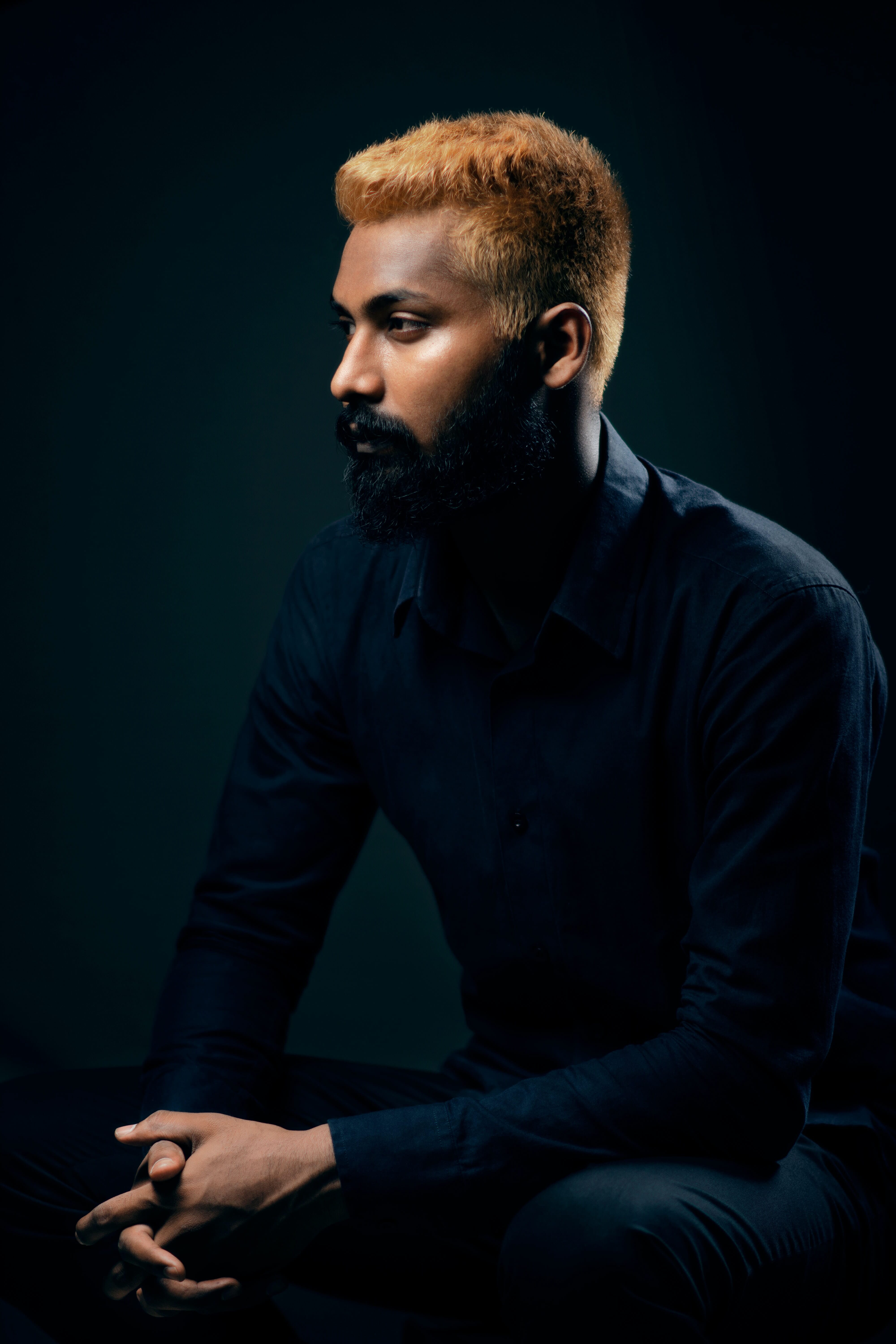 Man with blue clothes and a red shirt sitting dramatically in front of a dark backdrop.