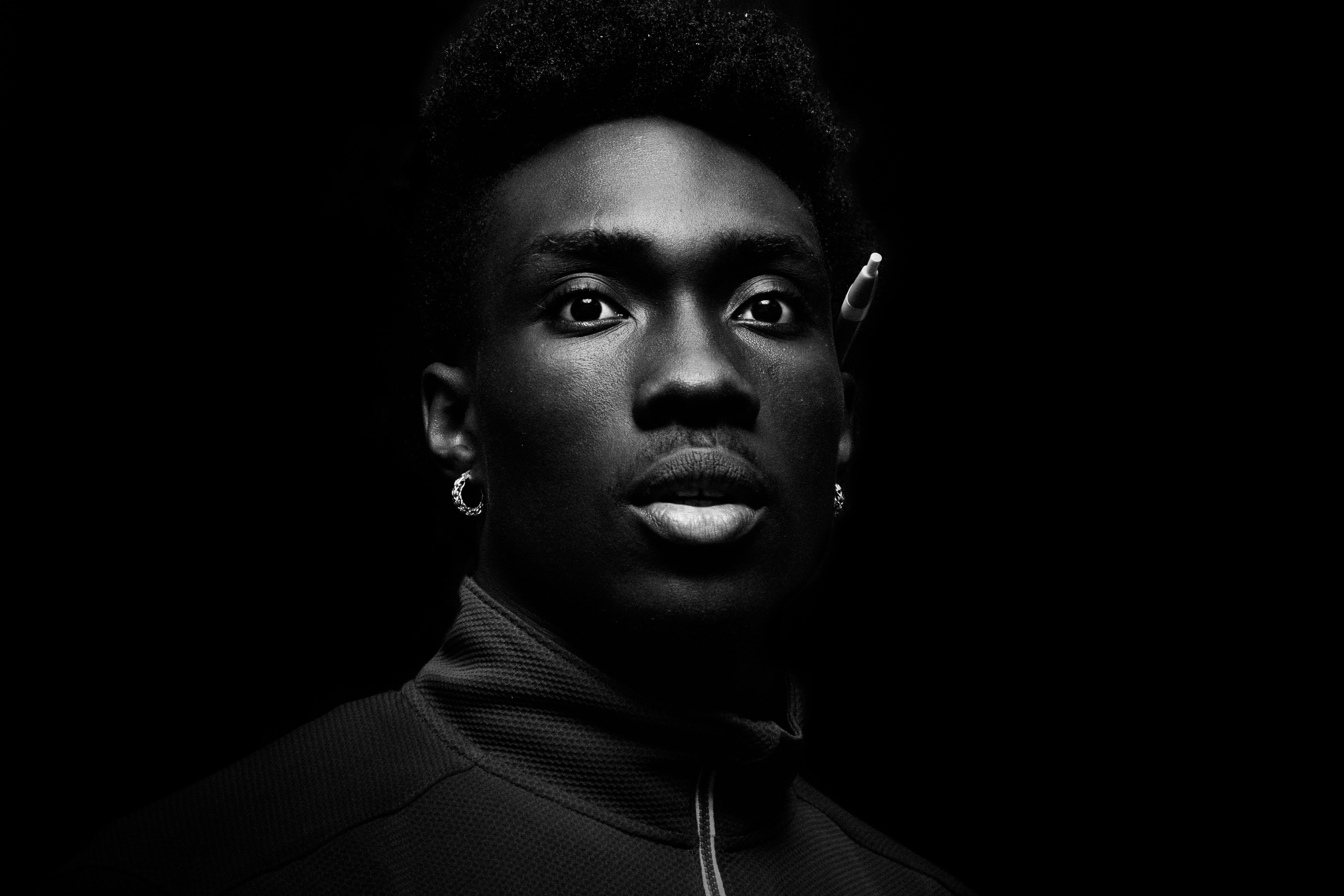 A striking portrait features a young man with dark skin, dramatic lighting accentuating his facial features against a black.