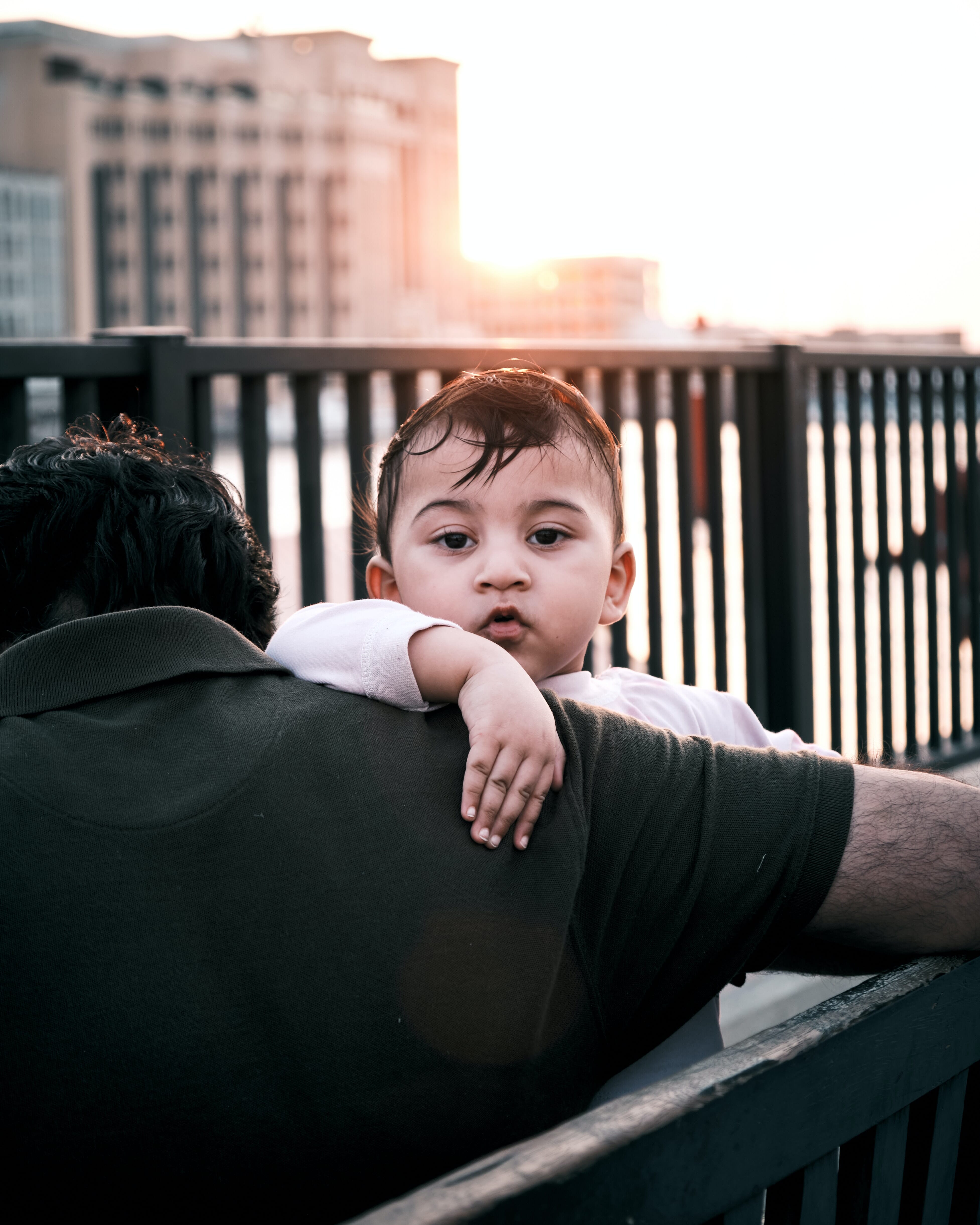 A child resting on a parent's shoulder is illuminated by warm sunset light. The background features a blurred cityscape.