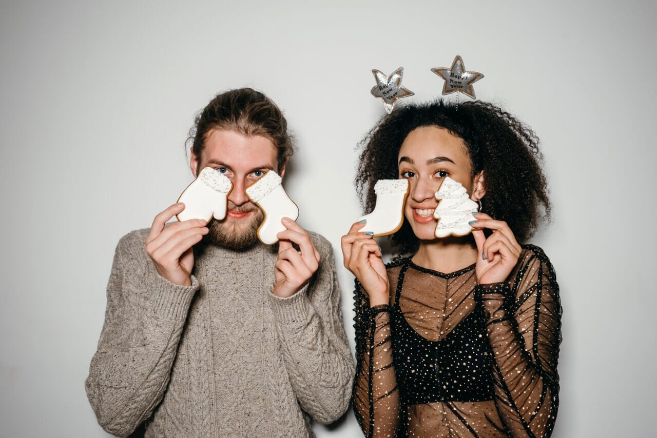 A man and a woman holding white Christmas cookies in front of their faces while standing before a white wall.