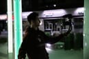 Man standing outside at night in urban setting, holding a camera with lavalier microphone filming himself between green lighted posts.