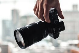 Hand holding a Sony a7 III with a black 24-70mm lens attached in front of a blurry background.