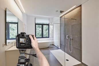 An outstretched hand on a DSLR camera mounted on a tripod capturing a photo of bathroom with a shower, a sink, and a bath.