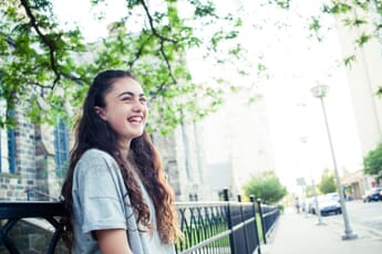 Girl leaning against a fence with a church and a leafy tree branch in the background.