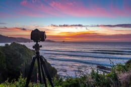 Camera on a tripod on a grassy hill overlooking the ocean and a blue, pink, and orange sunset sky.