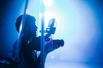 Man filming with a video camera that includes an external monitor in a room lit by bright blue lights.