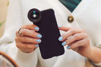 Person's hands with blue nails holding a smartphone with an external lens attached.
